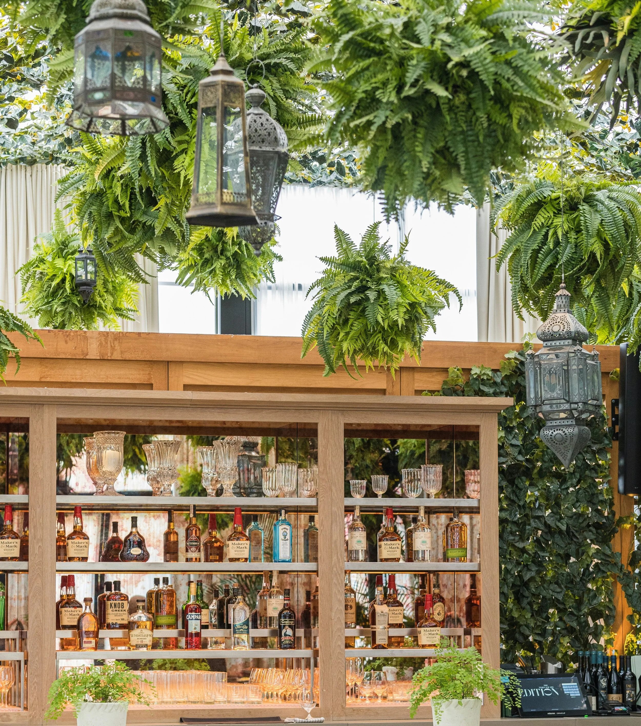 Indoor bar with wooden shelves displaying various liquor bottles and empty glasses, decorated with hanging lanterns and potted ferns, with bar equipment and bottles on the counter.