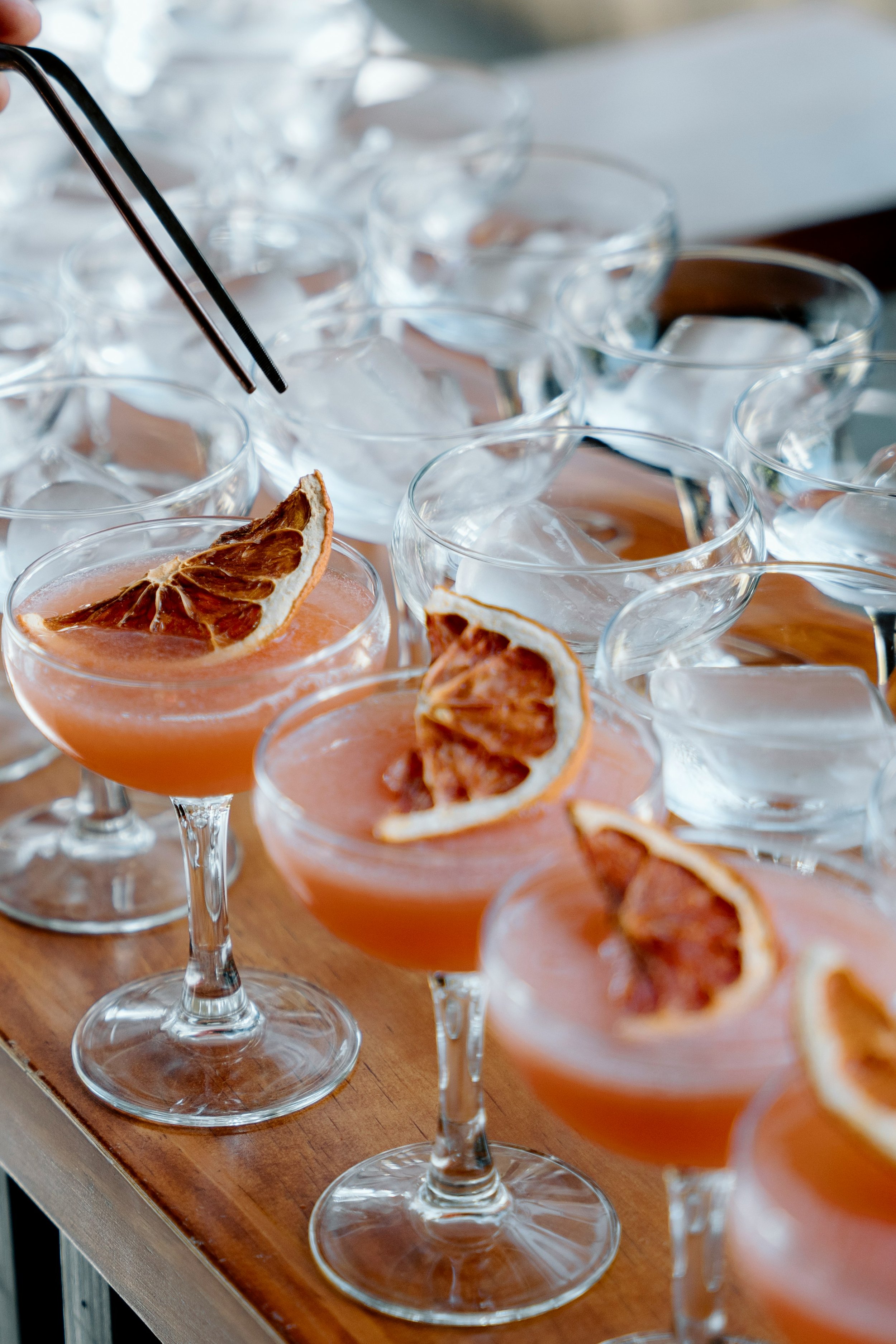 Multiple cocktail glasses with orange-colored drinks garnished with dried orange slices, arranged on a wooden surface.
