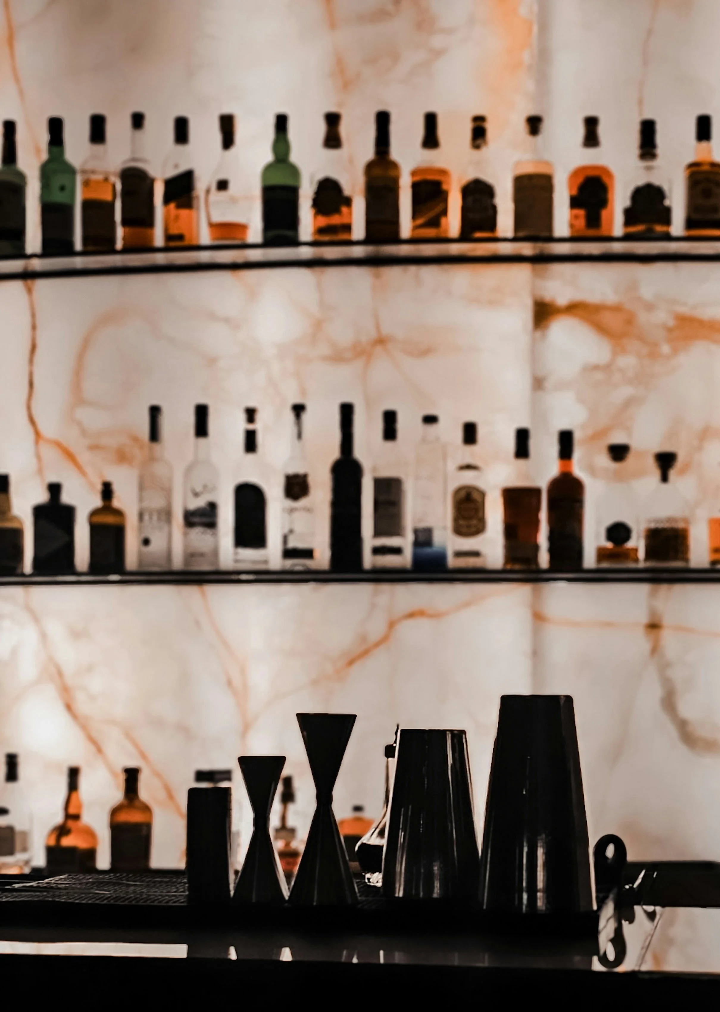 A bar shelf with a marble background, displaying various bottles of alcohol, with some bottles seen in the foreground and others arranged on the shelves behind.
