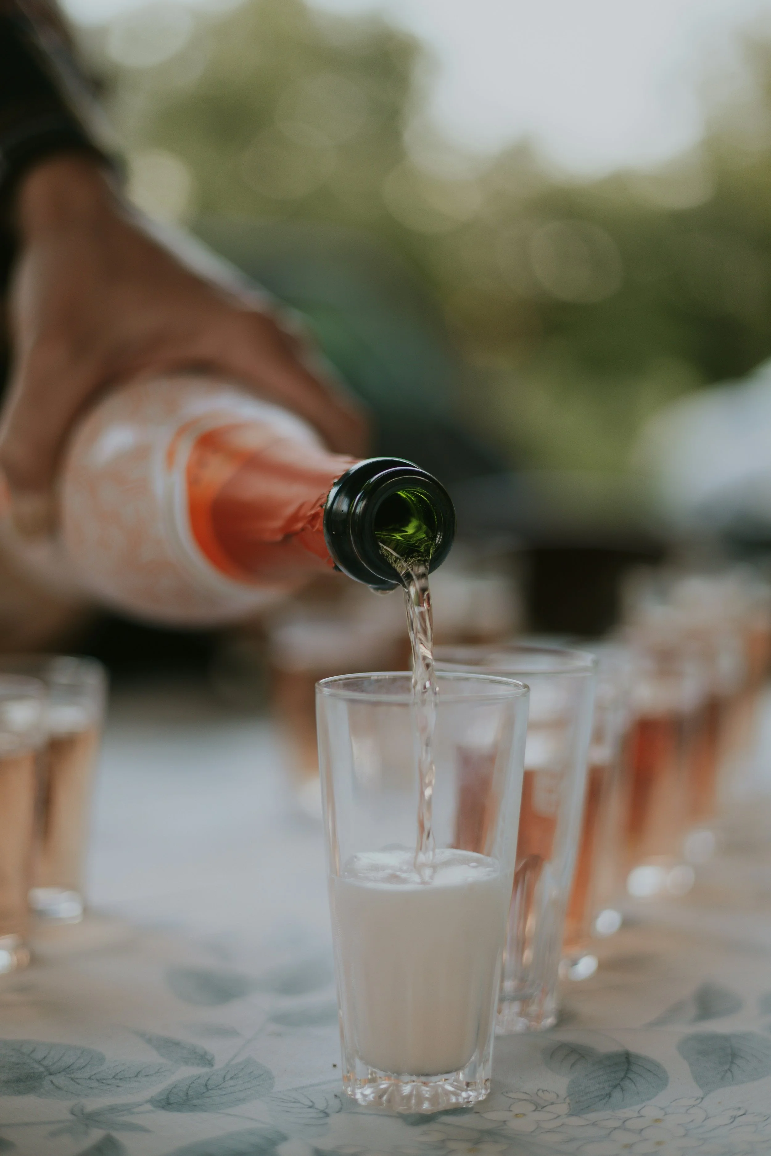 A person pouring a drink from a bottle into a clear glass, with several other glasses in the background, on a table with a floral tablecloth.