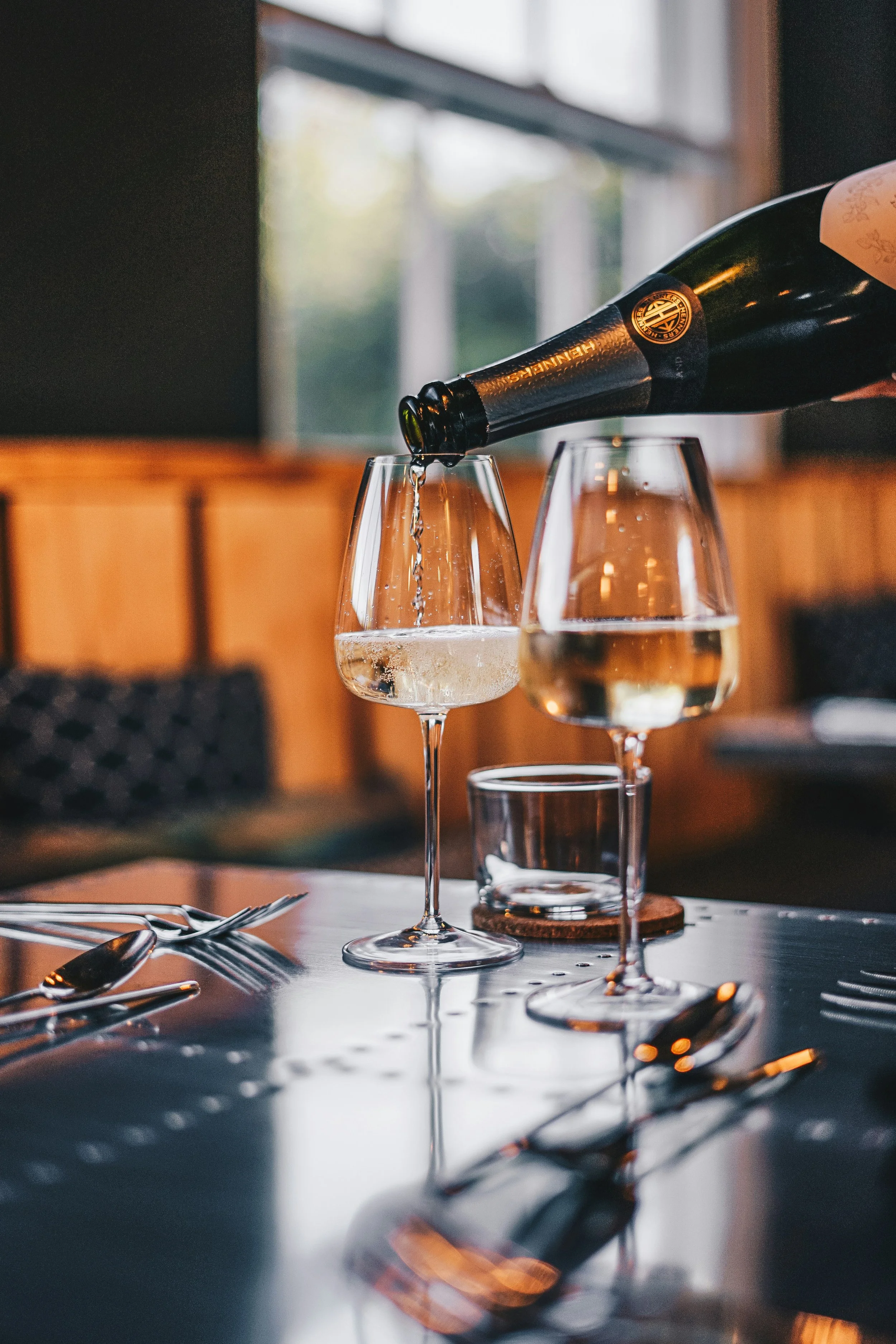 A wine bottle is pouring wine into a glass on a table with cutlery, in a cozy restaurant setting with natural light coming through a window.