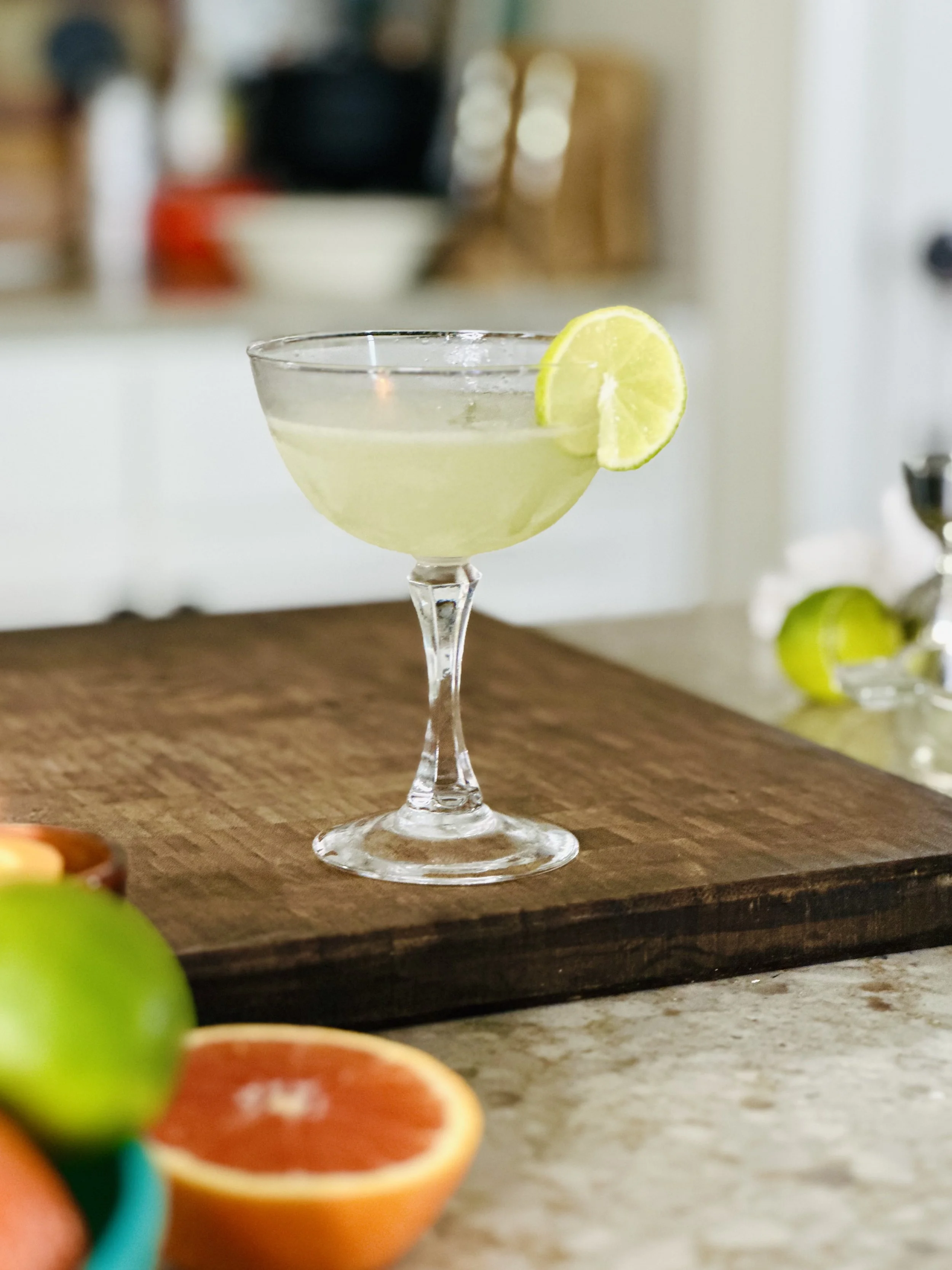 A cocktail glass with a lemon wheel garnish on the rim, filled with a pale yellow drink, placed on a wooden surface with colorful fruit in the foreground and a blurred kitchen background.