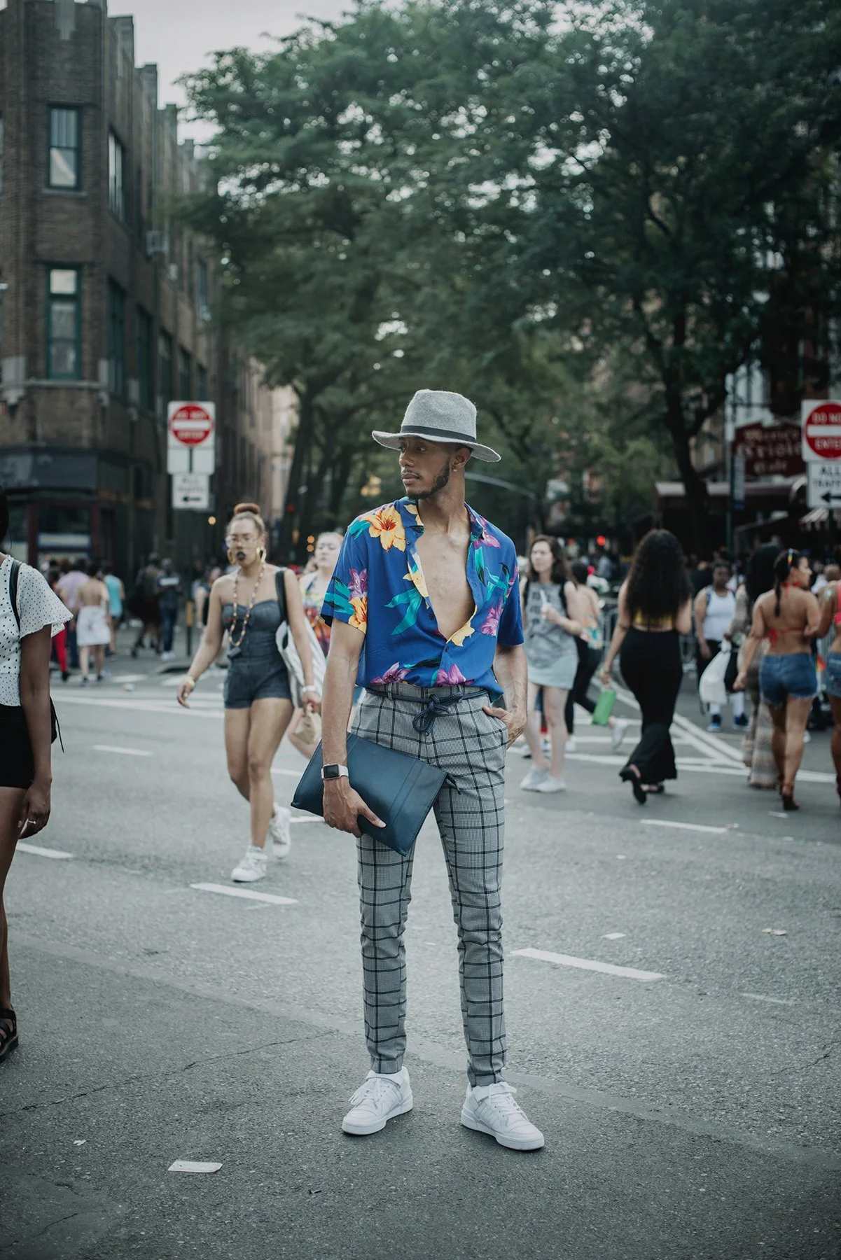 A young man standing in the middle of a busy city street, wearing a colorful tropical shirt, plaid pants, a gray wide-brimmed hat, and white sneakers, holding a black folder, with people walking behind him.