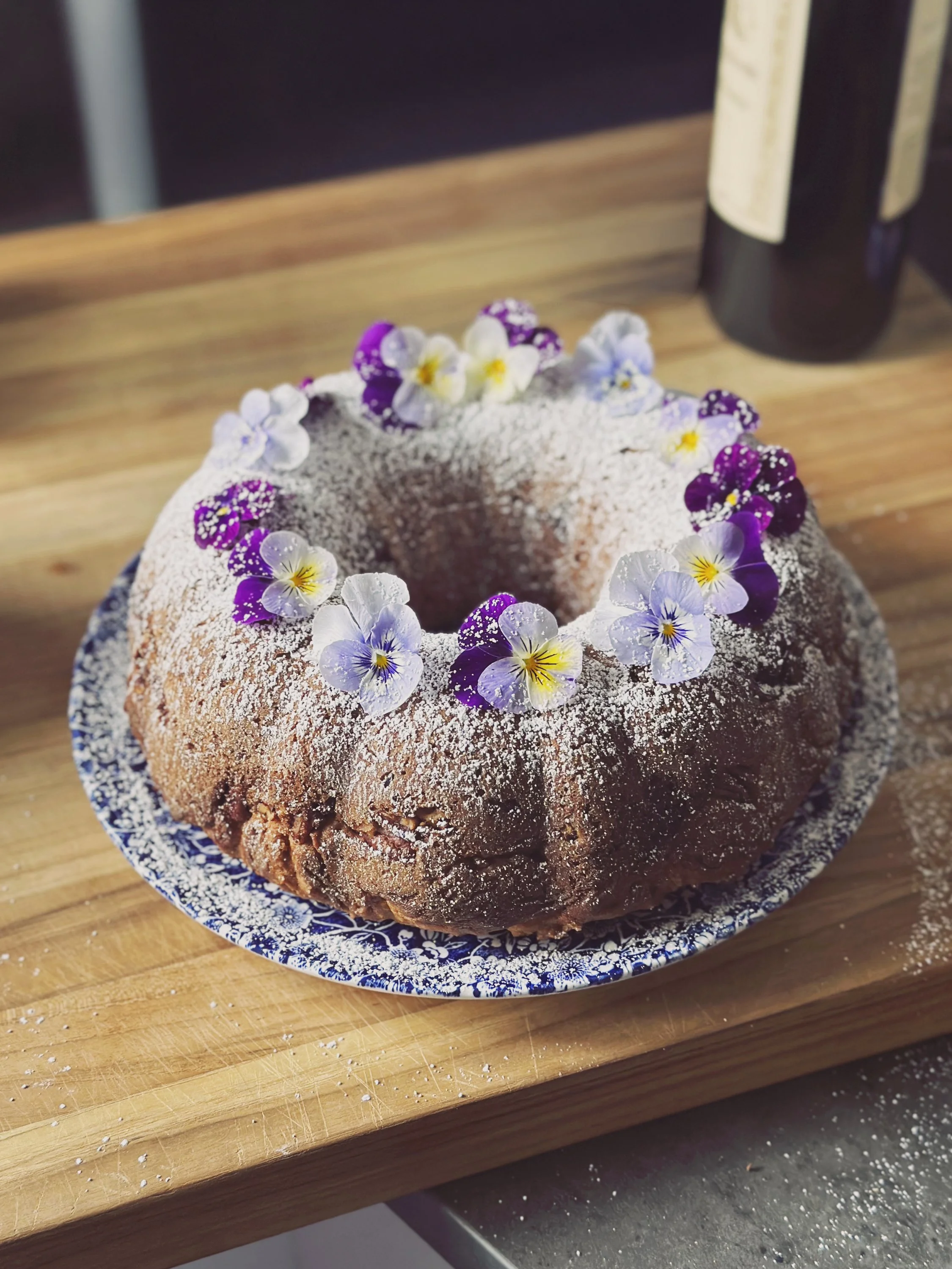 A bundt cake decorated with edible flowers and powdered sugar on a blue and white plate placed on a wooden surface.