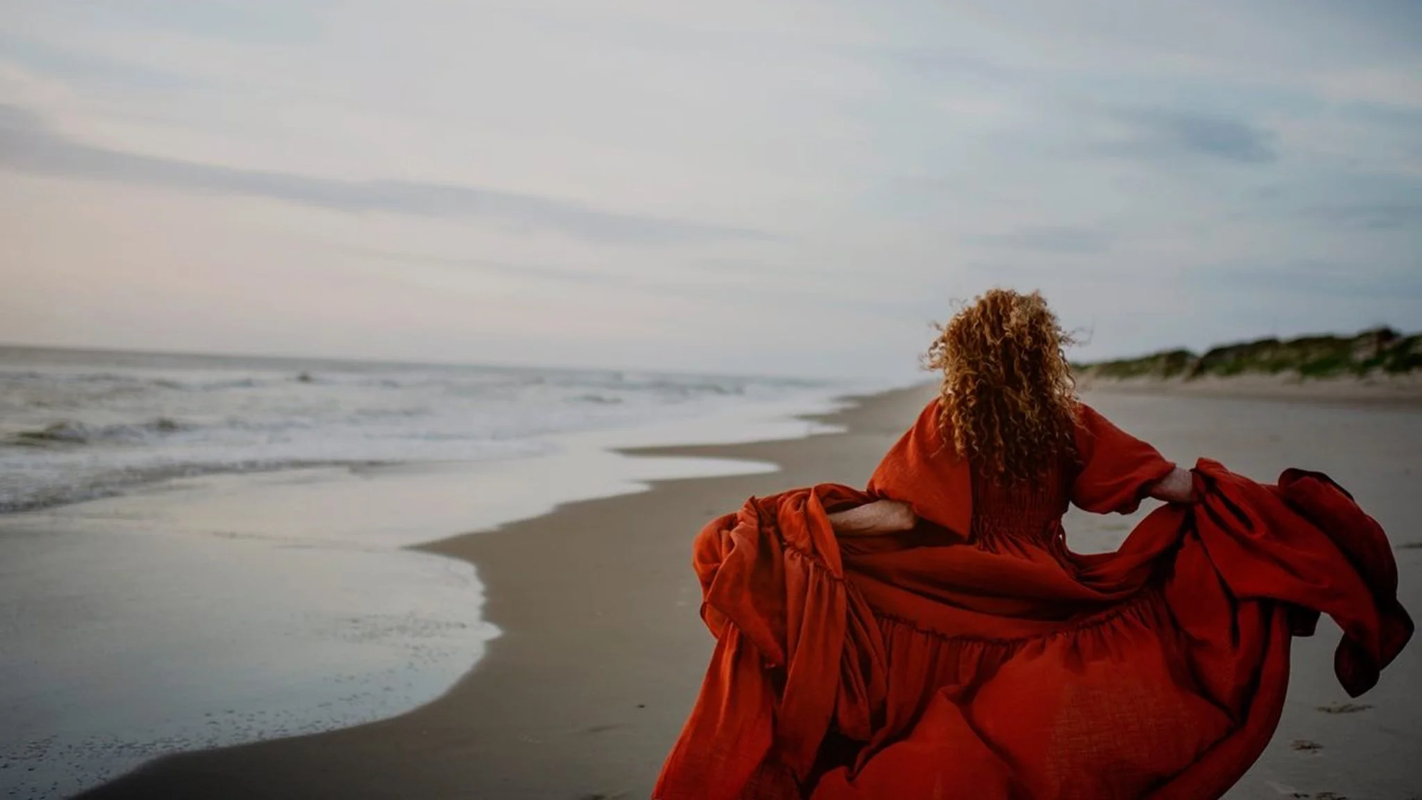 A woman with curly red hair, wearing a long red dress, sitting on the beach and looking toward the ocean with a cloudy sky.