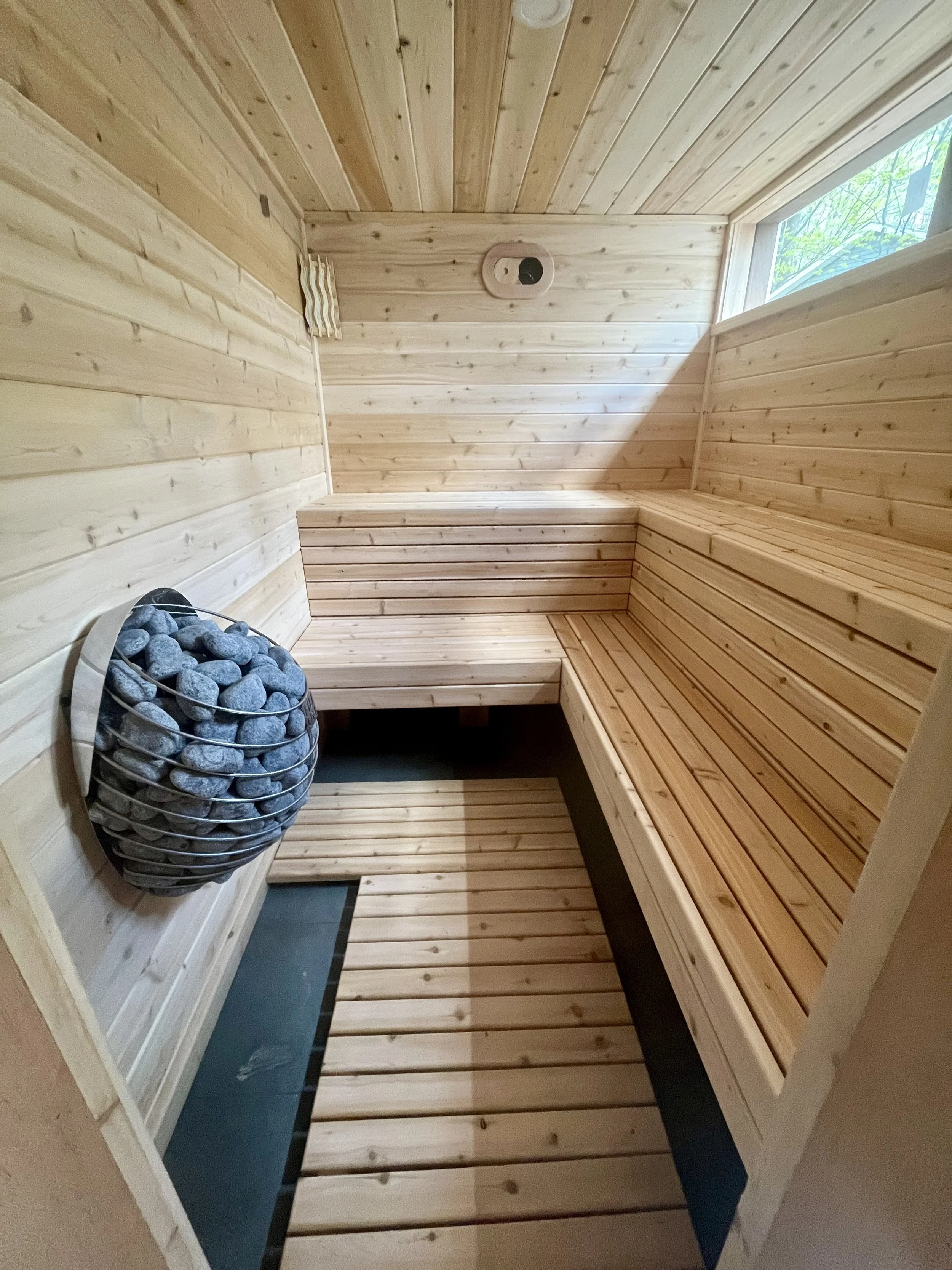A custom cedar sauna room with benches, a window, and a heater with rocks.