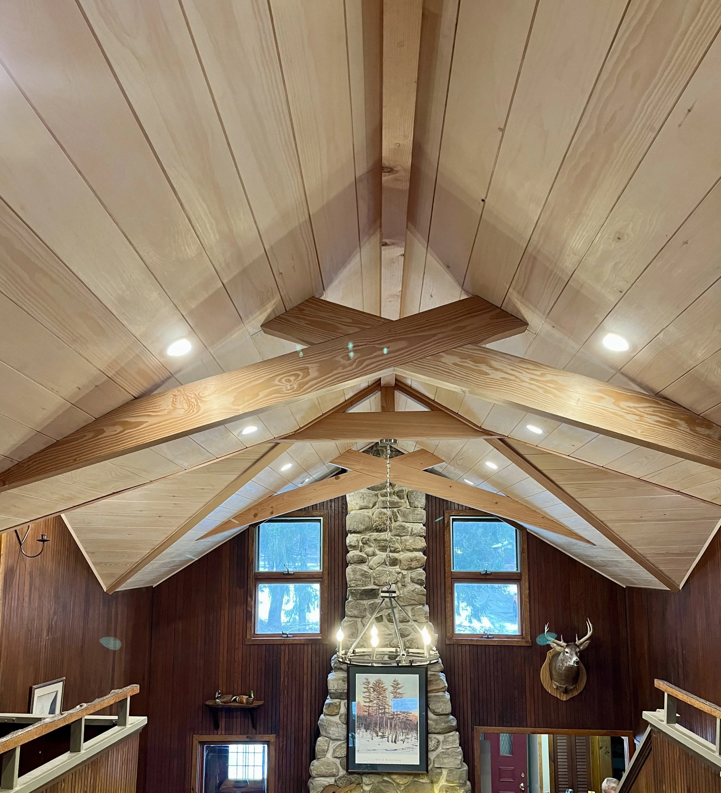 Interior of a rustic lodge or cabin with a high wooden ceiling, custom scissor trusses, stone fireplace, large windows, and mounted animal head.