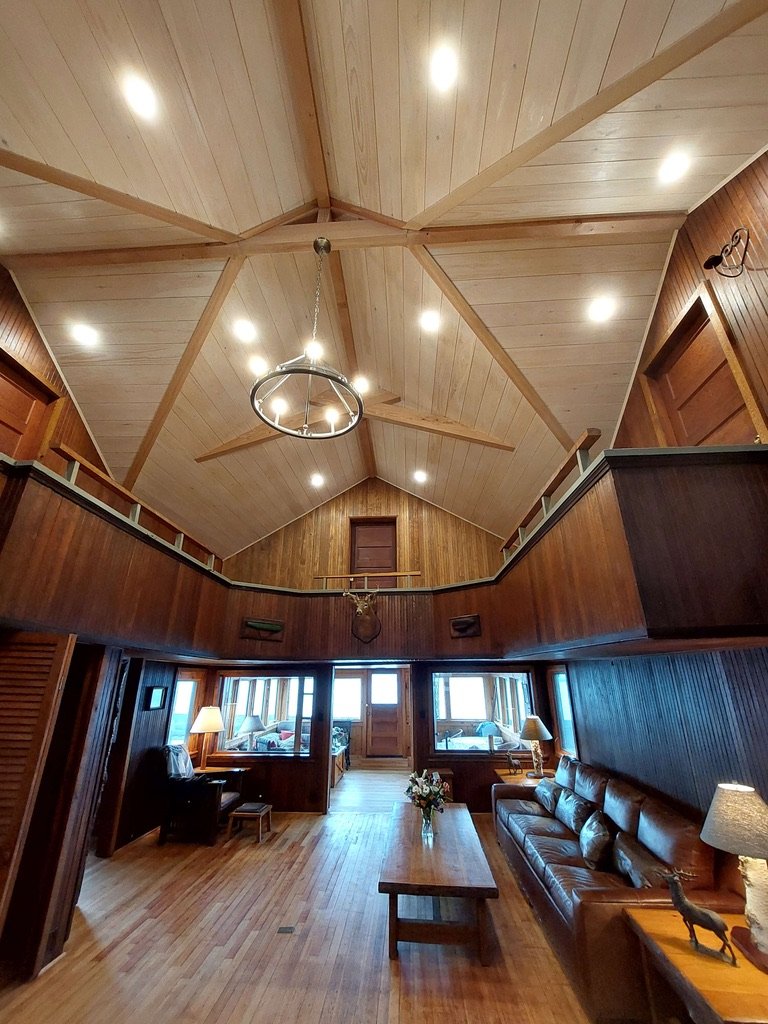 Interior of a spacious wooden cabin living room with high vaulted ceiling, wooden walls, and hardwood floor. Large window area with seating, coffee table, and side lamps. Ceiling lights and a chandelier are visible.