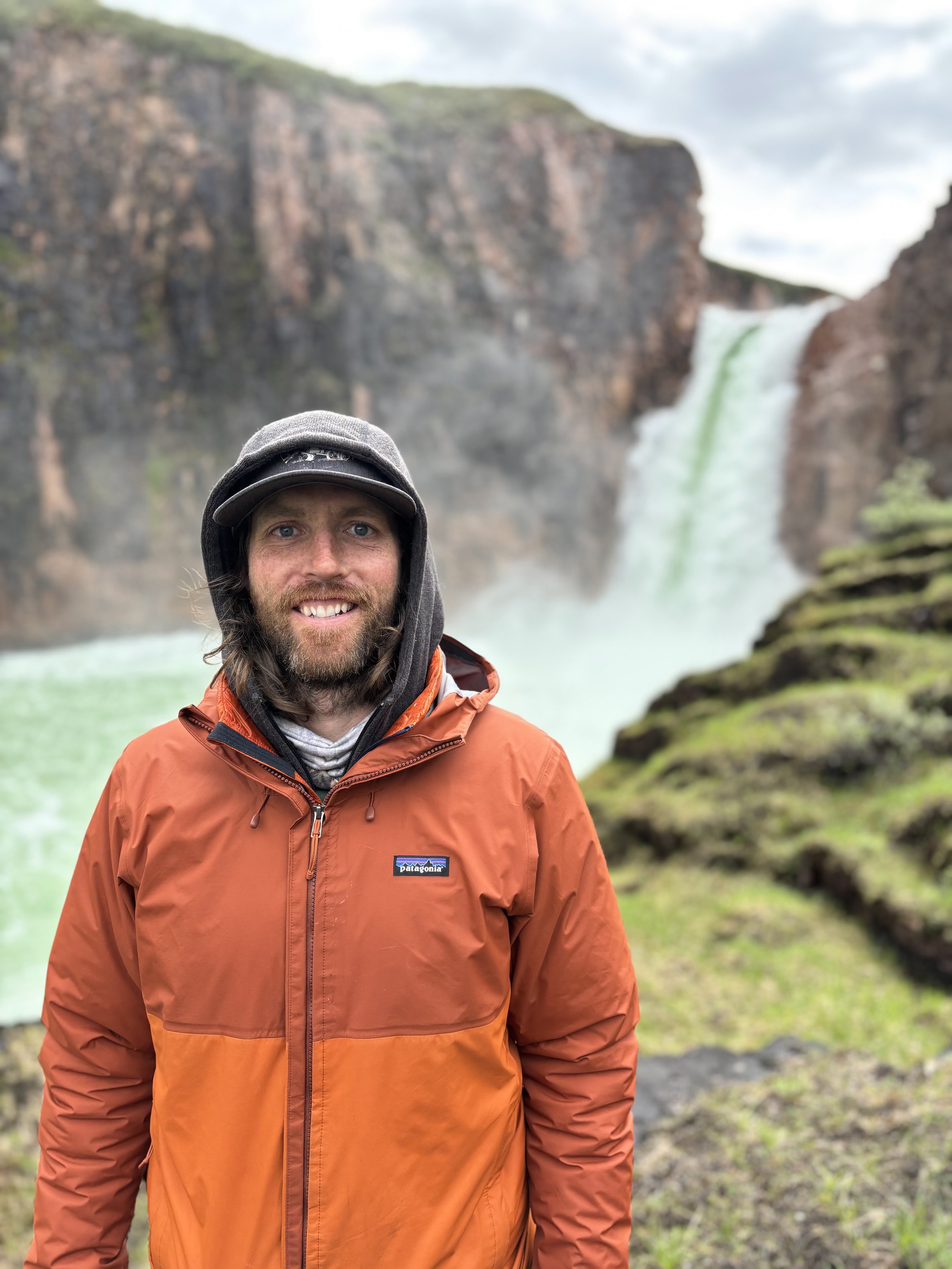 A man smiling and wearing an orange Patagonia jacket, a gray hoodie, and a gray cap standing outdoors near a large waterfall surrounded by green moss-covered rocks and mist, with a cloudy sky in the background.