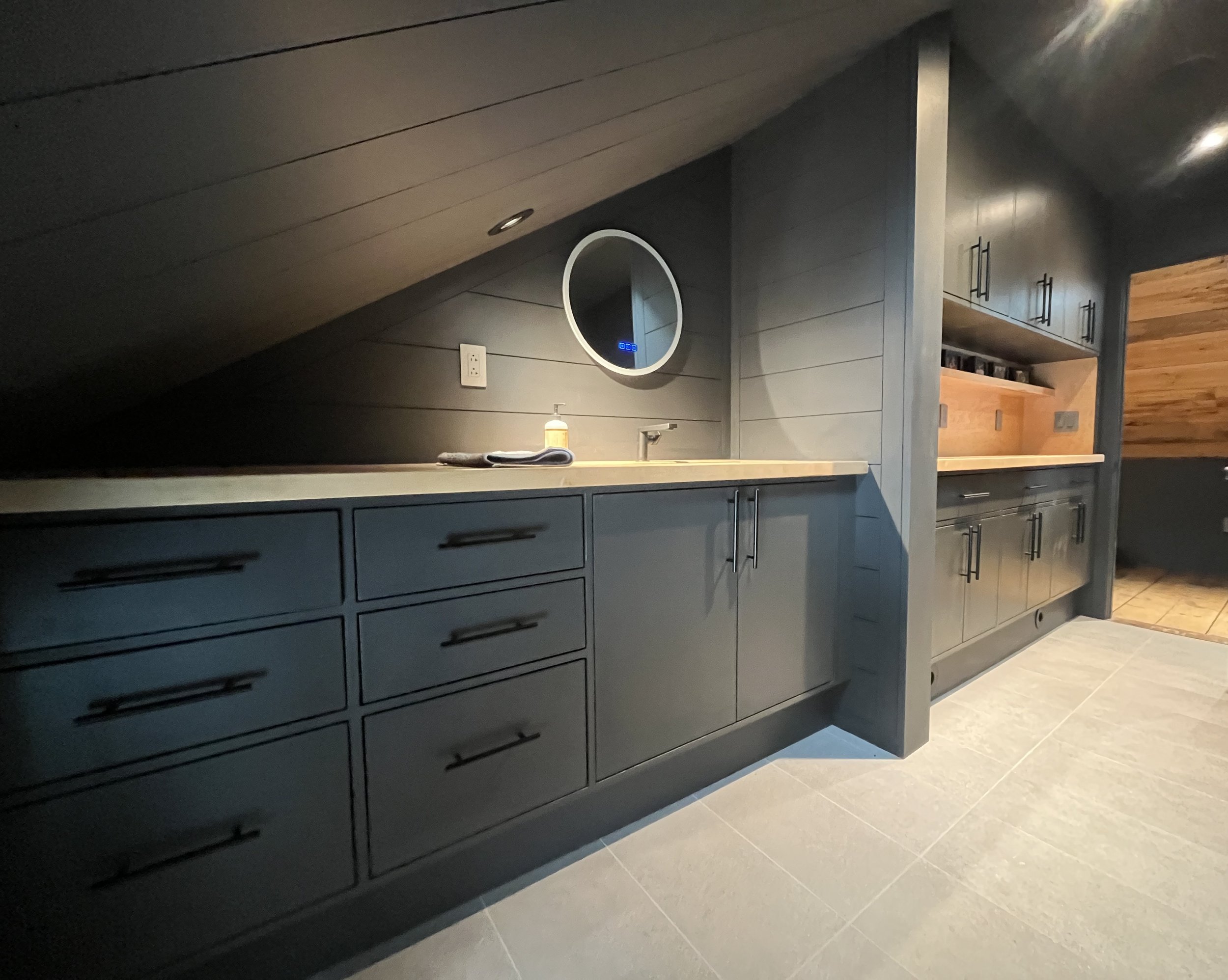 Modern kitchen with gray cabinets, a light-colored countertop, and a circular mirror on the wall. There is a bottle and a folded towel on the countertop, with wood accents and tile flooring visible.