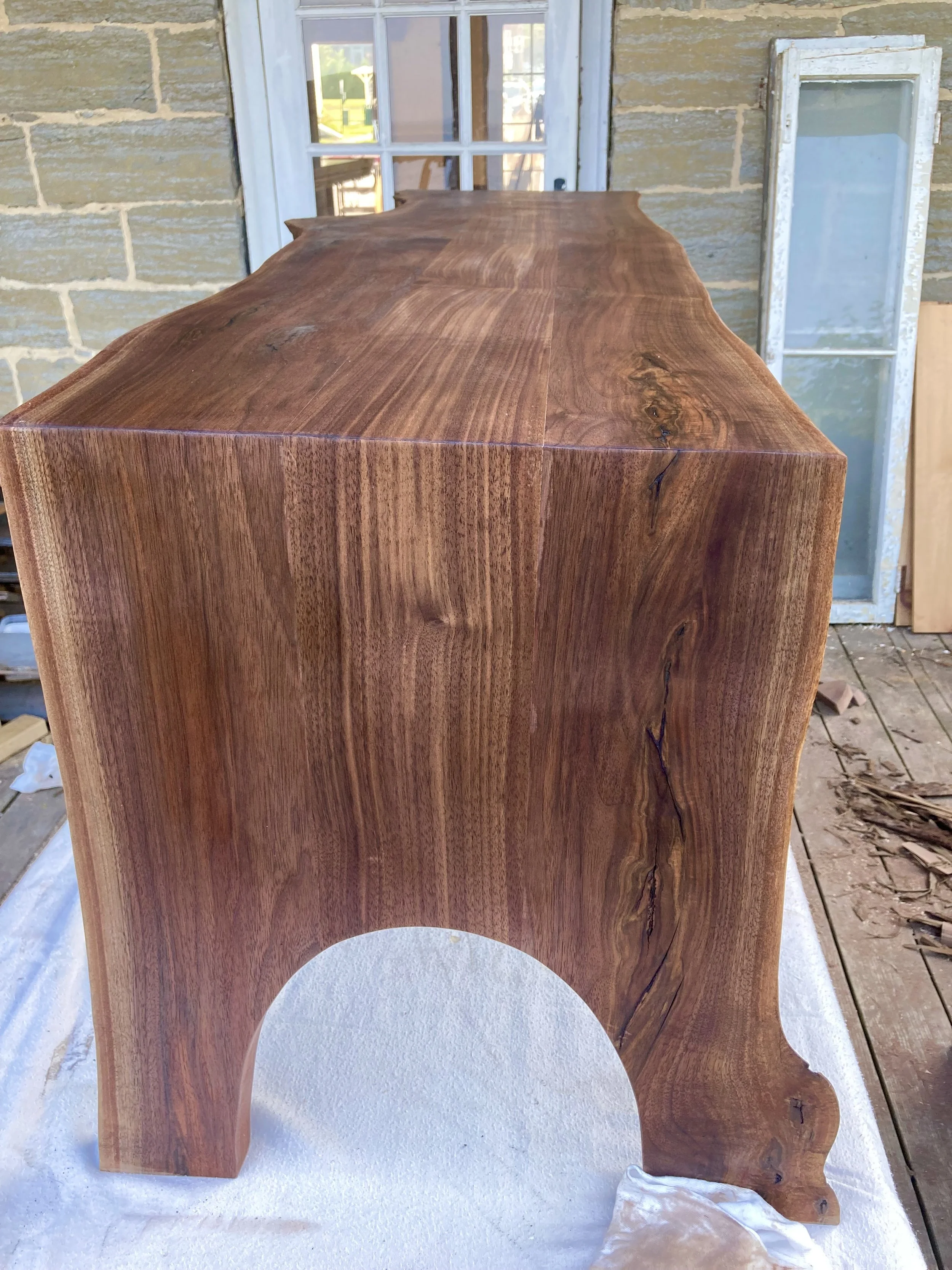 A large, polished walnut waterfall table with natural curves, positioned outdoors against a brick wall with a window and a white window frame in the background.