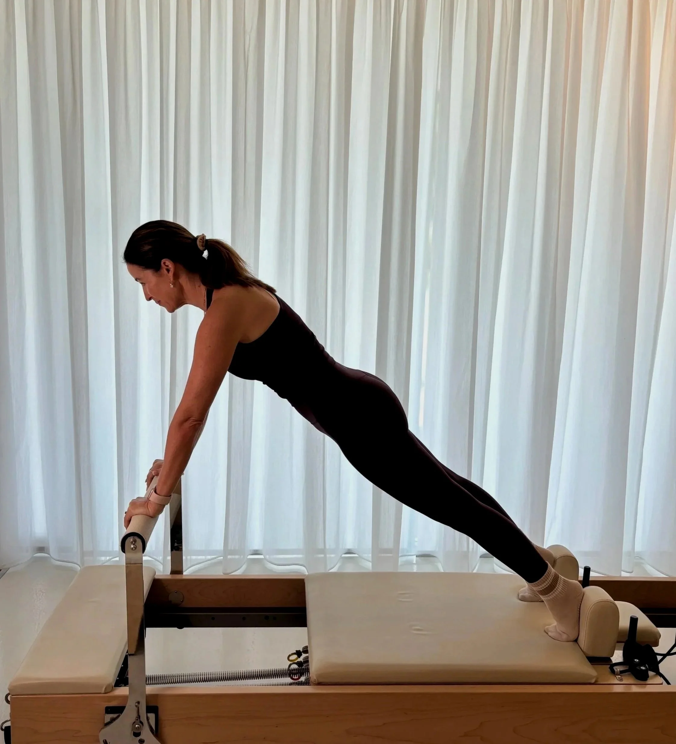 Woman in black clothes practicing yoga or stretching on Reformer Pilates machine in a studio with white curtains.