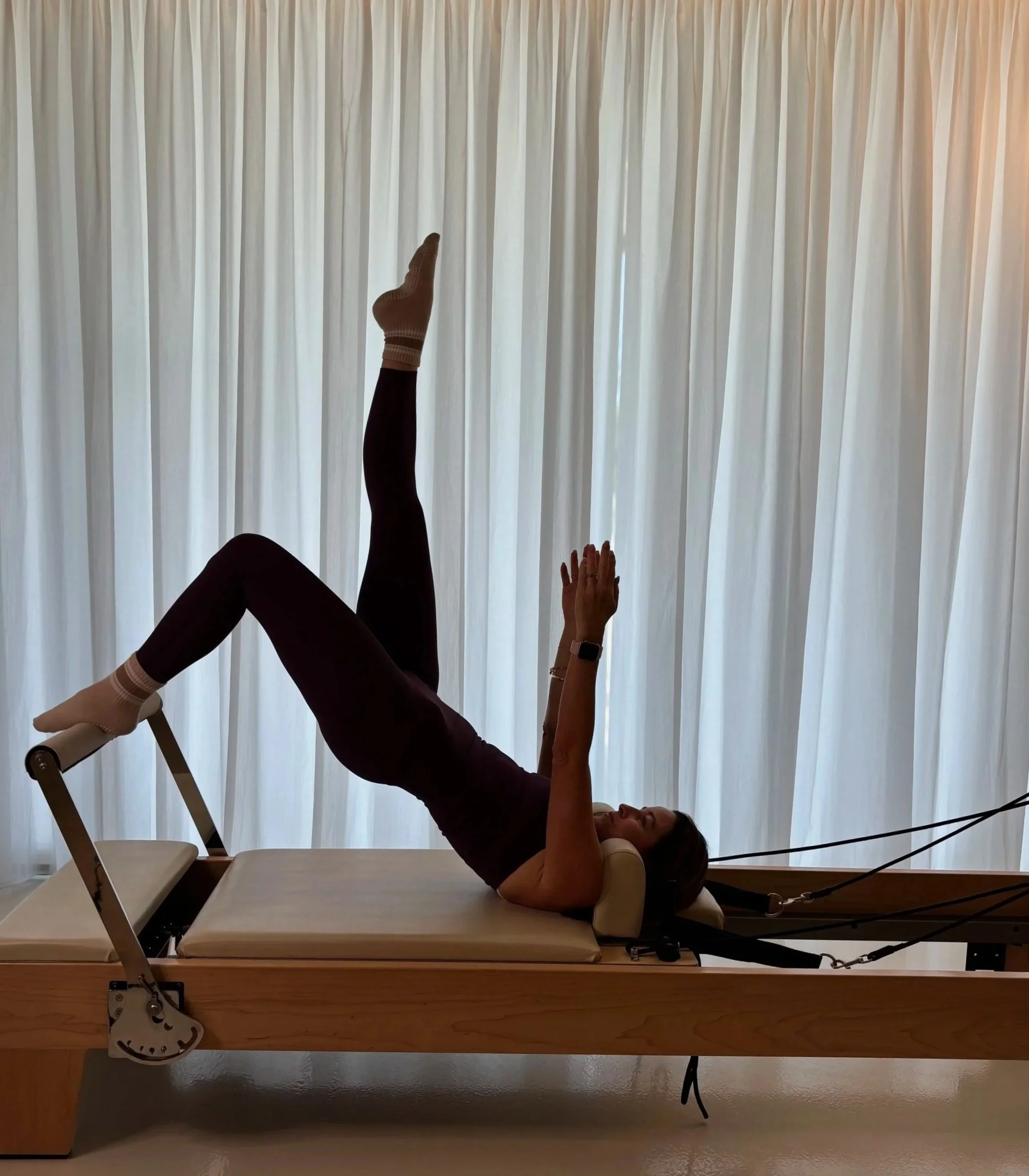A woman lying on an examination table with her legs raised, wearing socks and a wristwatch, in a room with light-colored curtains.