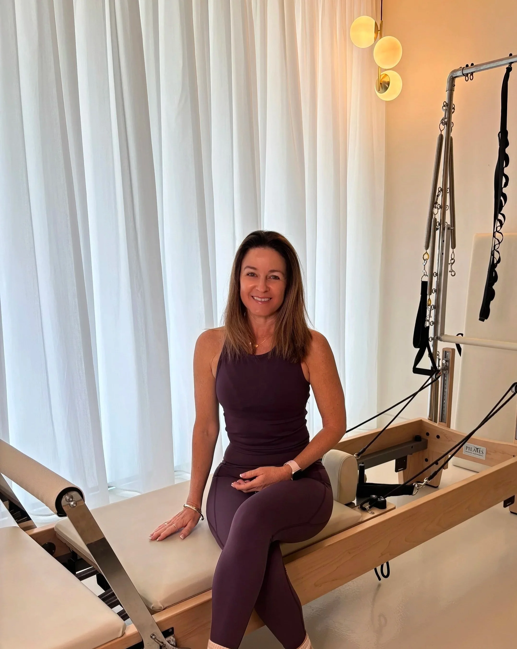 A woman sitting on a Pilates reformer machine in a room with white curtains and modern lighting.