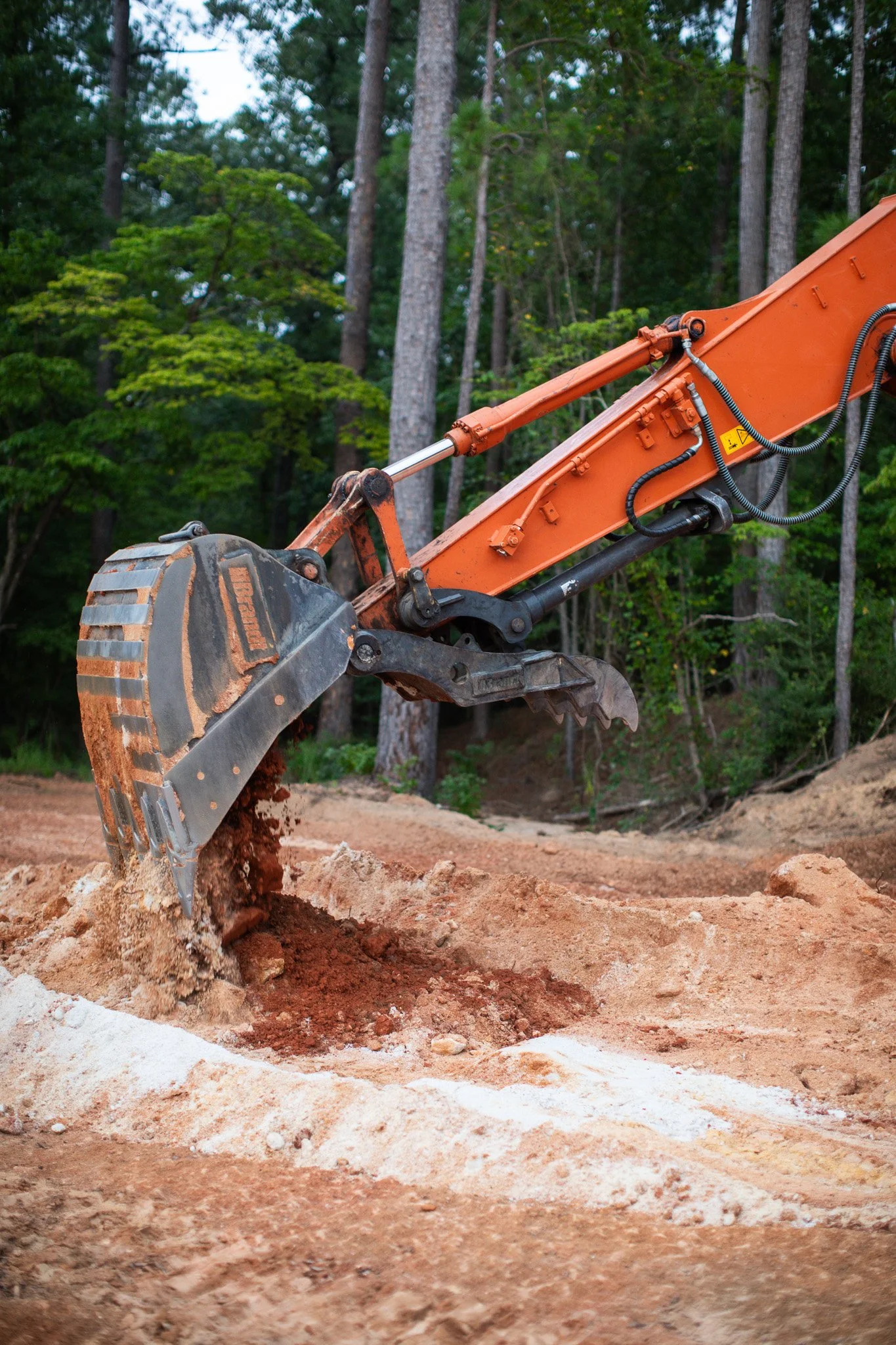 An orange construction excavator digging into reddish-brown soil in a forest clearing, with tall trees and green foliage in the background.