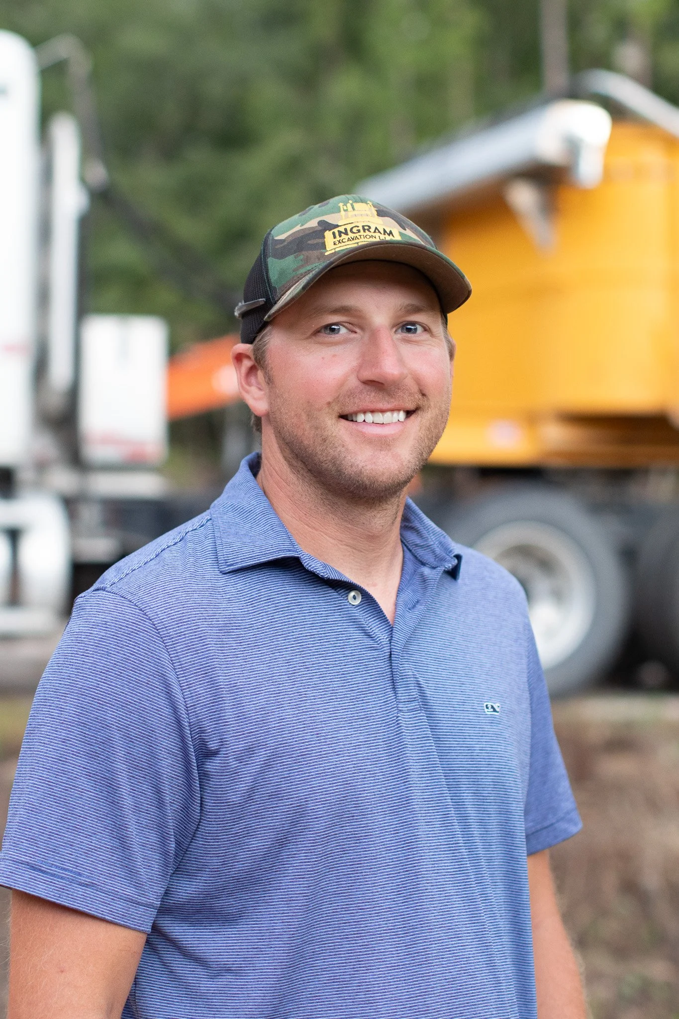 A man wearing a camouflage cap with the logo 'Ingram Excavation LLC' and a blue striped polo shirt, smiling outdoors with blurry trees and construction equipment in the background.