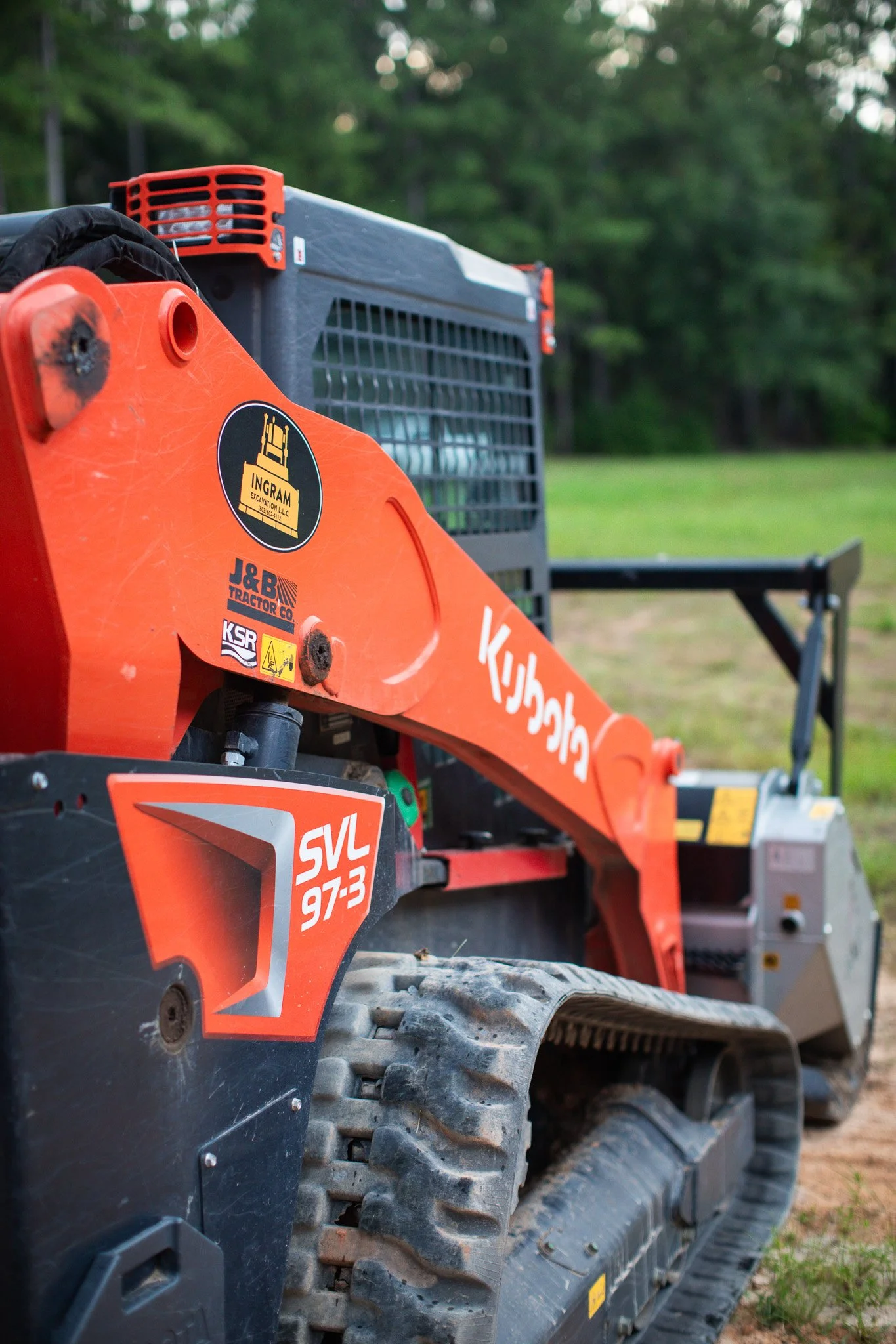 A close-up of an orange Kubota compact excavator, showing details of the tracks, side panel, and engine components, with a blurred background of green trees and grass.