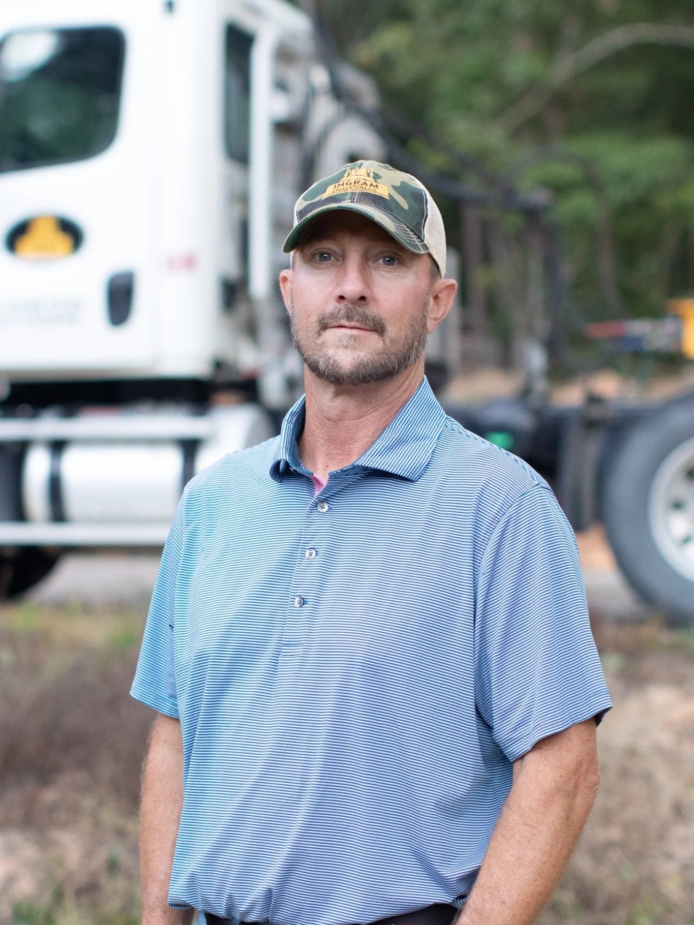 A man in a blue striped collared shirt and camo baseball cap stands outdoors with trees and a truck in the background.
