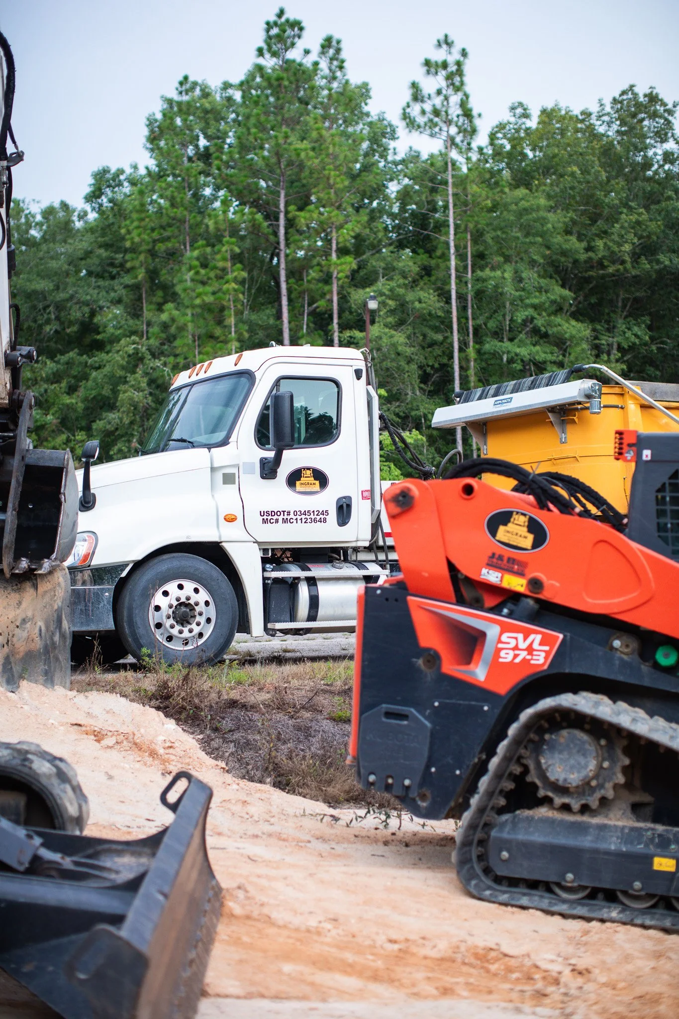 Construction site with construction equipment including a white truck, orange and black machinery, and a yellow container, with trees and a lamppost in the background.