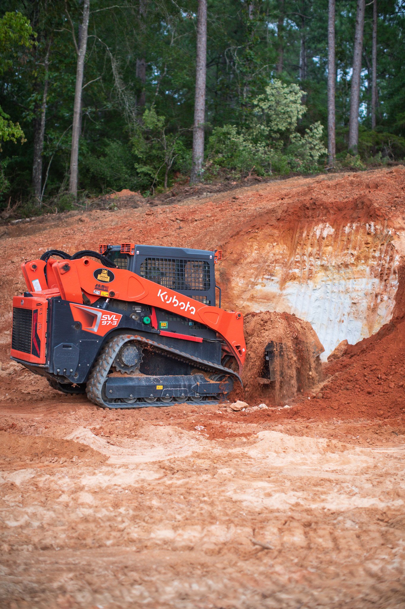 A small orange and black tracked excavator working on a dirt and clay slope with forest trees in the background.