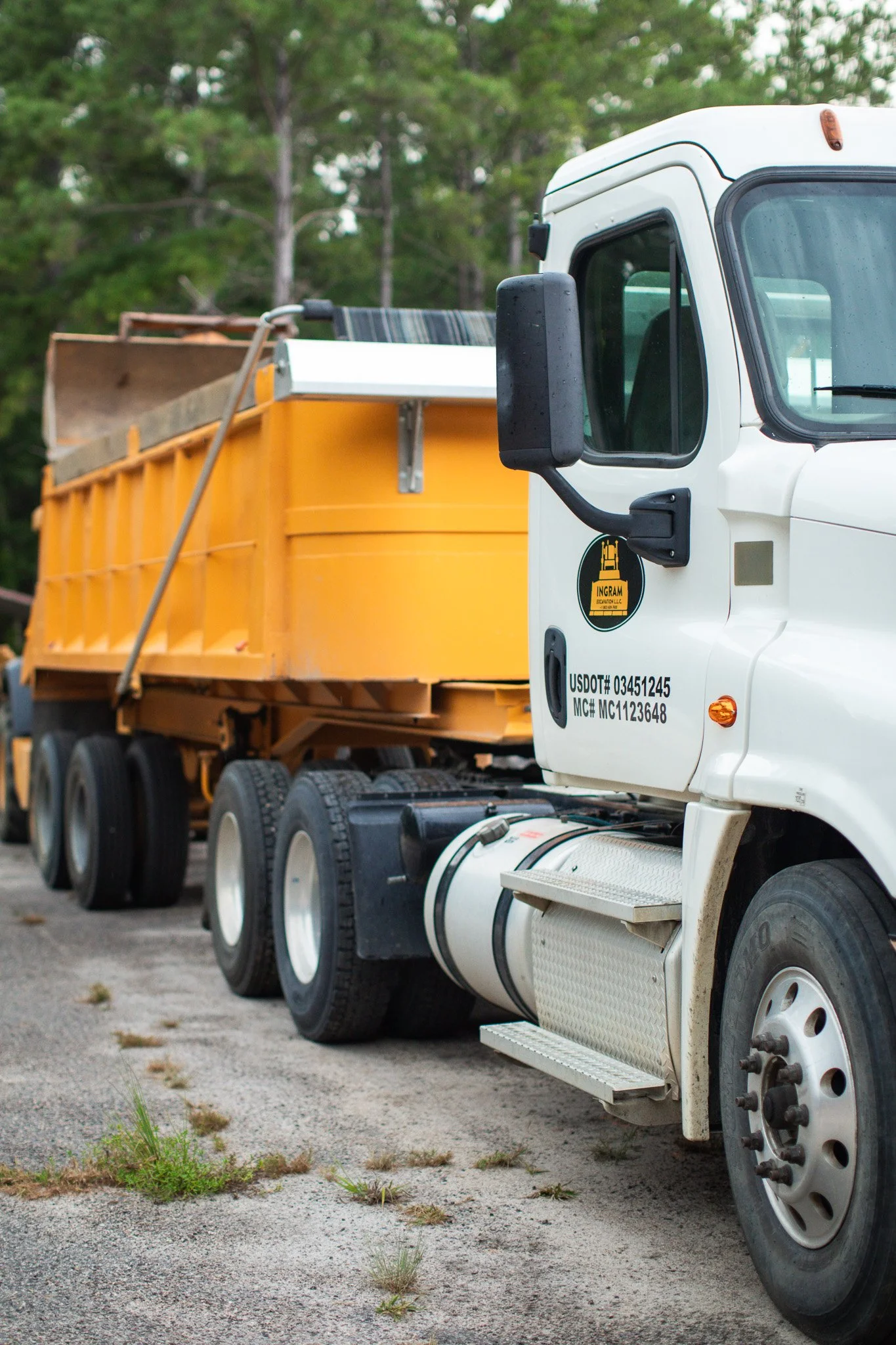 Close-up of a white dump truck with an orange bed parked on a gravel surface, with trees in the background.