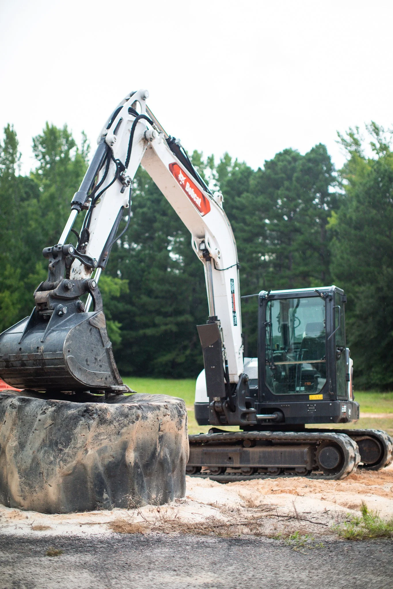 A construction excavator with a white body and black tracks parked on a dirt surface, with green trees in the background.