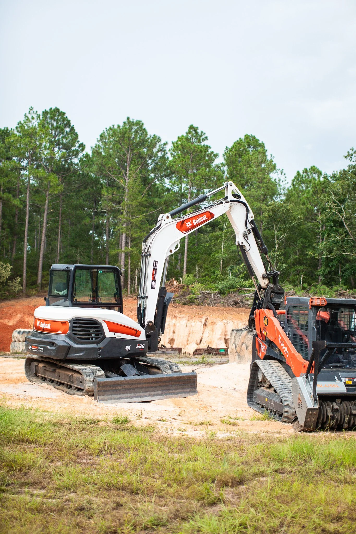 Construction site with two small excavators, one with a Bobcat logo, on dirt ground with grass and trees in the background.
