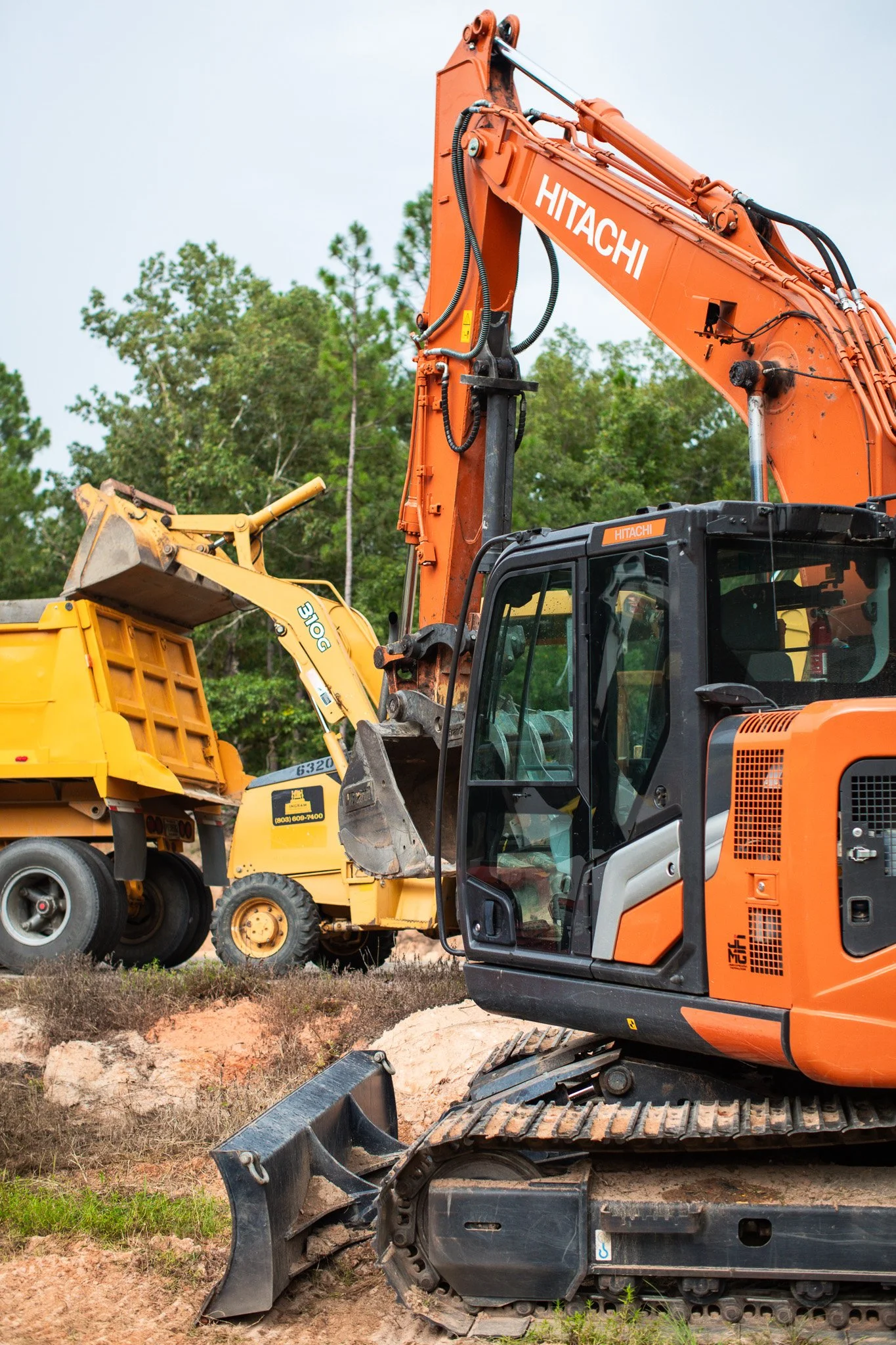 A construction site with orange and yellow heavy machinery, including an excavator with tracks, in front of green trees under a cloudy sky.