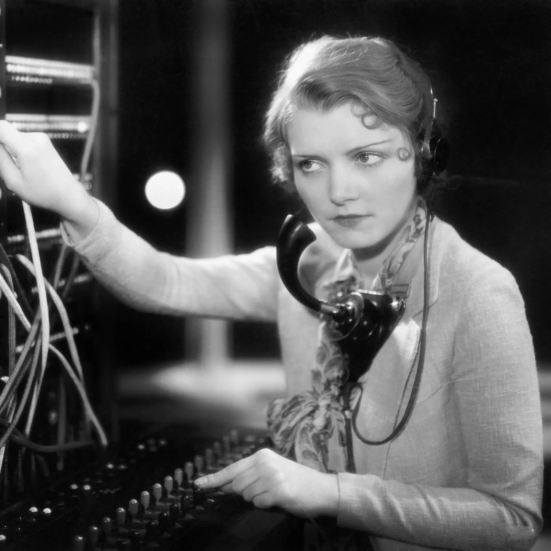 A photo of a telephone operator with blond hair, wearing headphones and a scarf, using an antique telephone switchboard