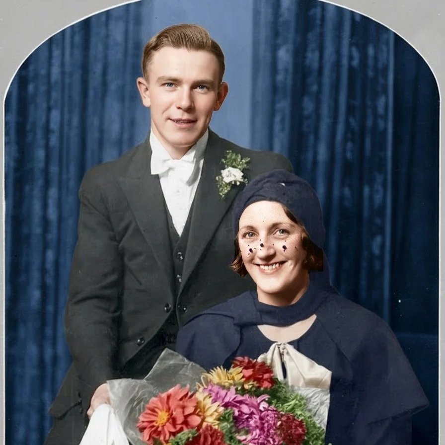 A young man in a suit and a young woman in a vintage costume are posing together for a portrait, with a dark blue curtain backdrop.
