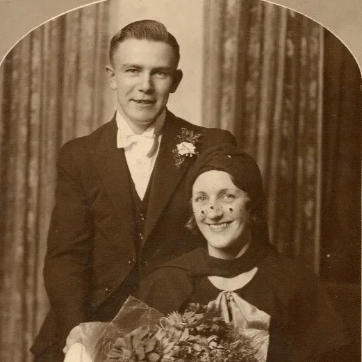 A vintage black-and-white photograph of a young man in a suit with a boutonniere and a young woman dressed as a clown with face paint, holding a bouquet of flowers, posing for a photo in front of a curtain.