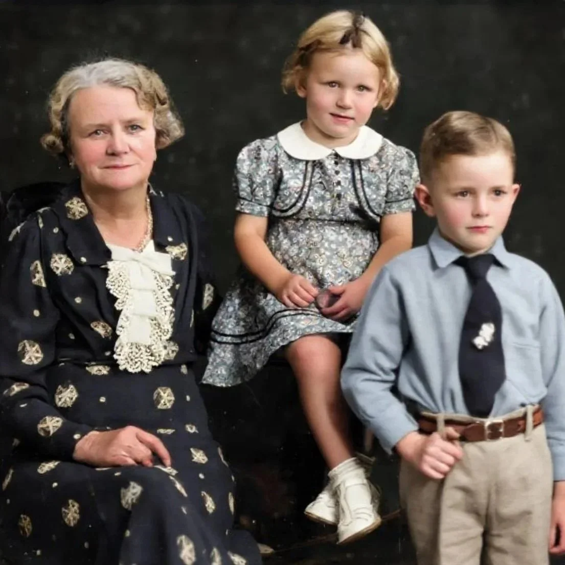 A colourised photo of an elderly woman with short grey hair and a young girl with blonde hair sitting beside a young boy with short brown hair, against a dark background.