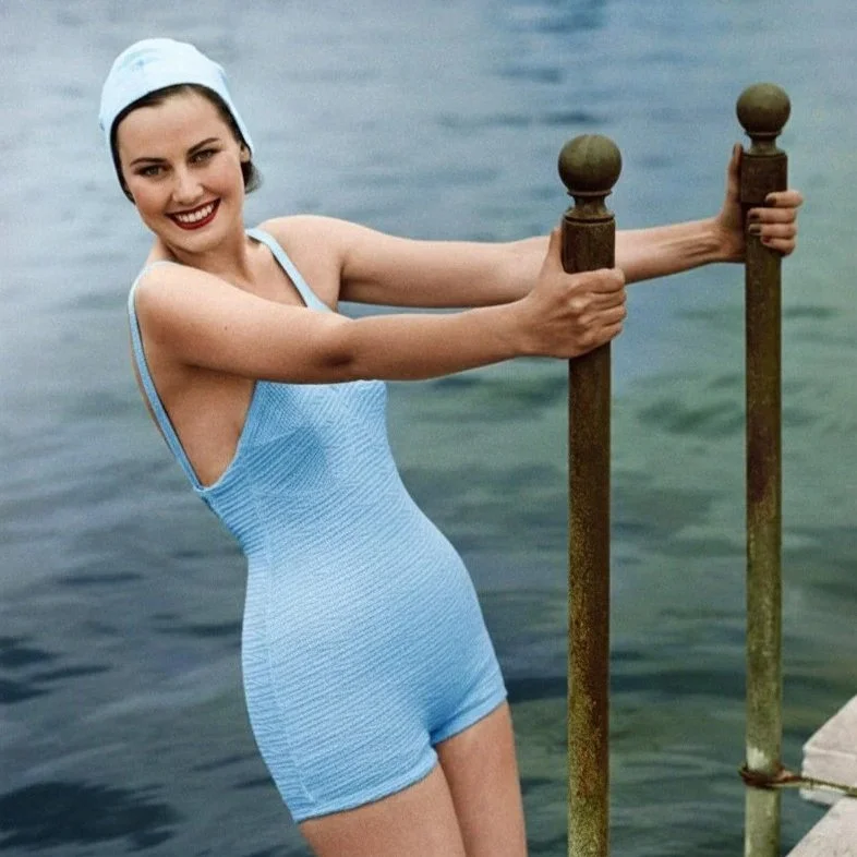 A woman in a light blue swimsuit and matching bathing cap standing by the water, holding onto a rusty metal railing, smiling at the camera.