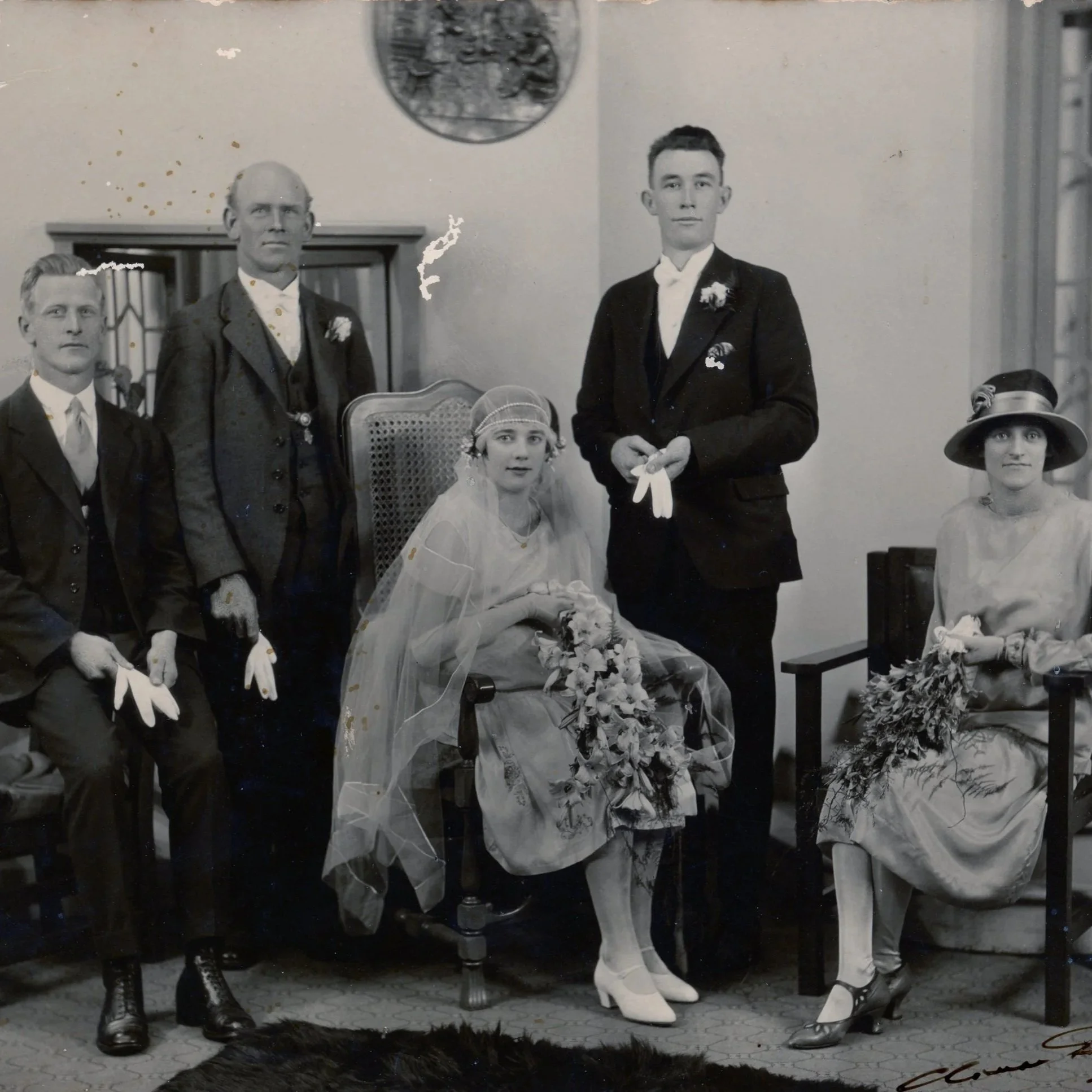 Black and white historical photograph of a wedding party with five people, including the bride and groom seated in the center. The bride wears a wedding dress and veil, holding a bouquet, while the groom wears a suit with a white bow tie and boutonni