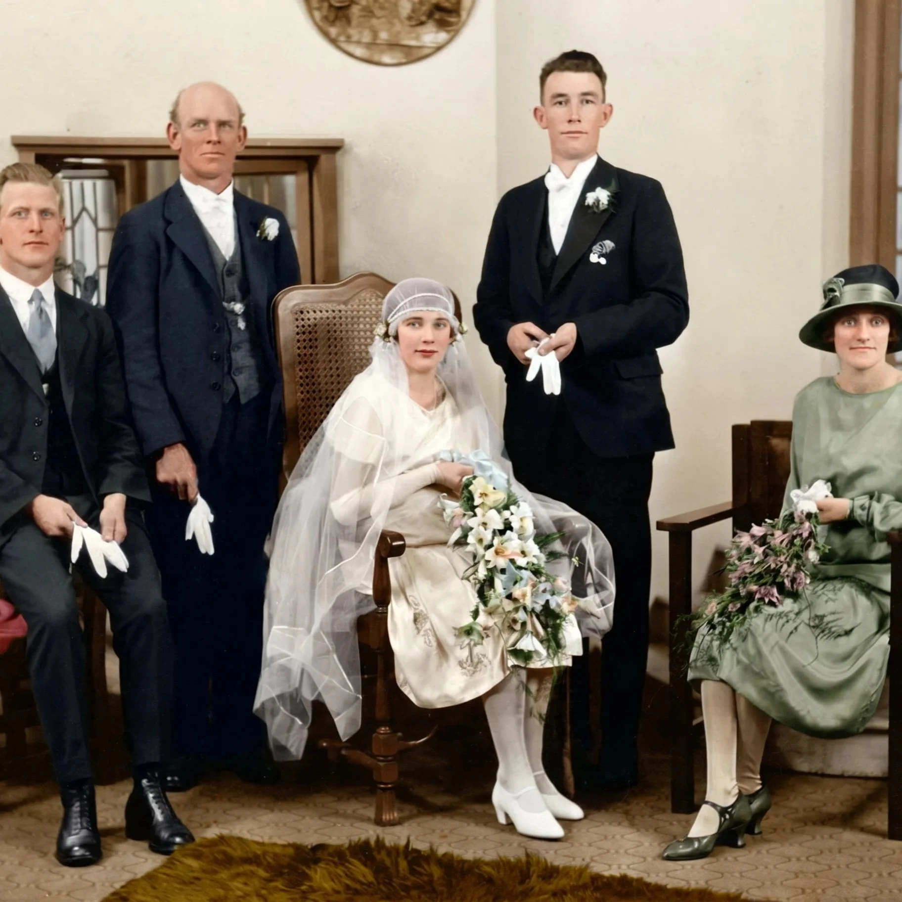 A vintage wedding scene with four men and one woman dressed in early 20th century attire. The woman, seated, wears a white wedding dress with a veil and holds a bouquet of white flowers. The men are in suits, with the groom in a black tuxedo with a w