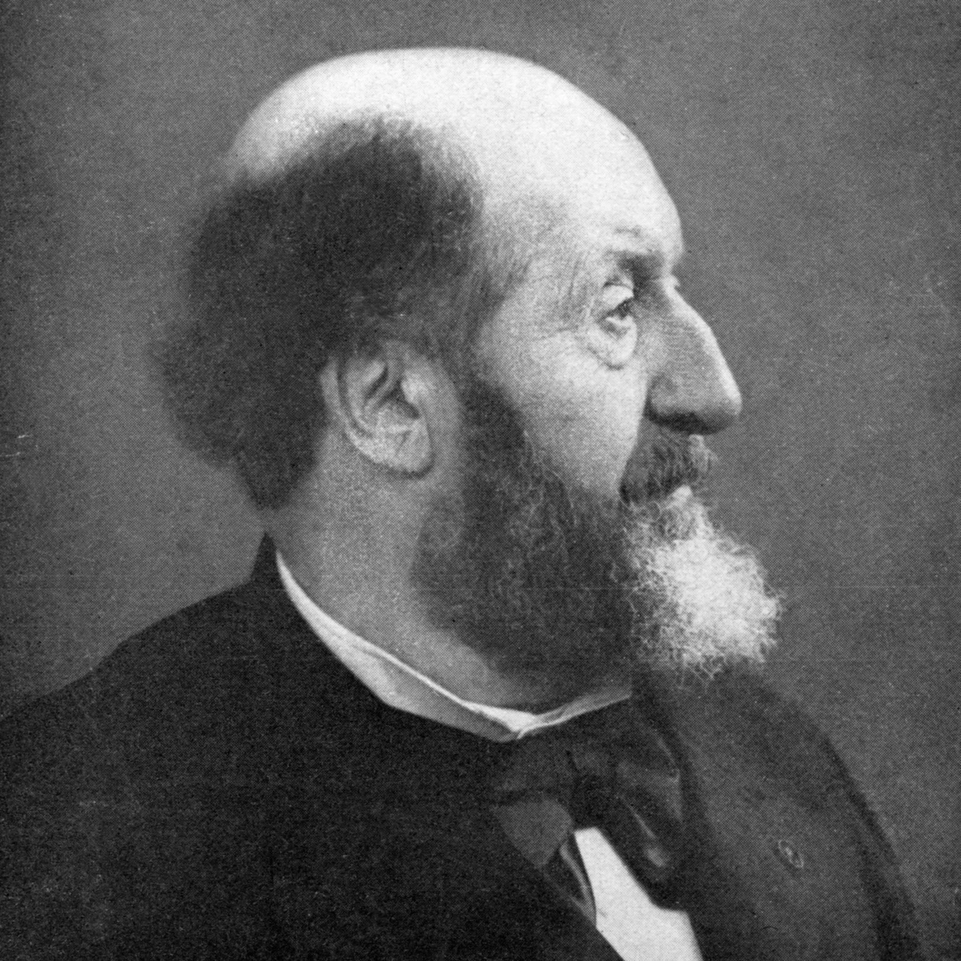 A black and white photograph of a man in profile, dressed in formal attire, with a high forehead, beard, and mustache, set against a plain background.