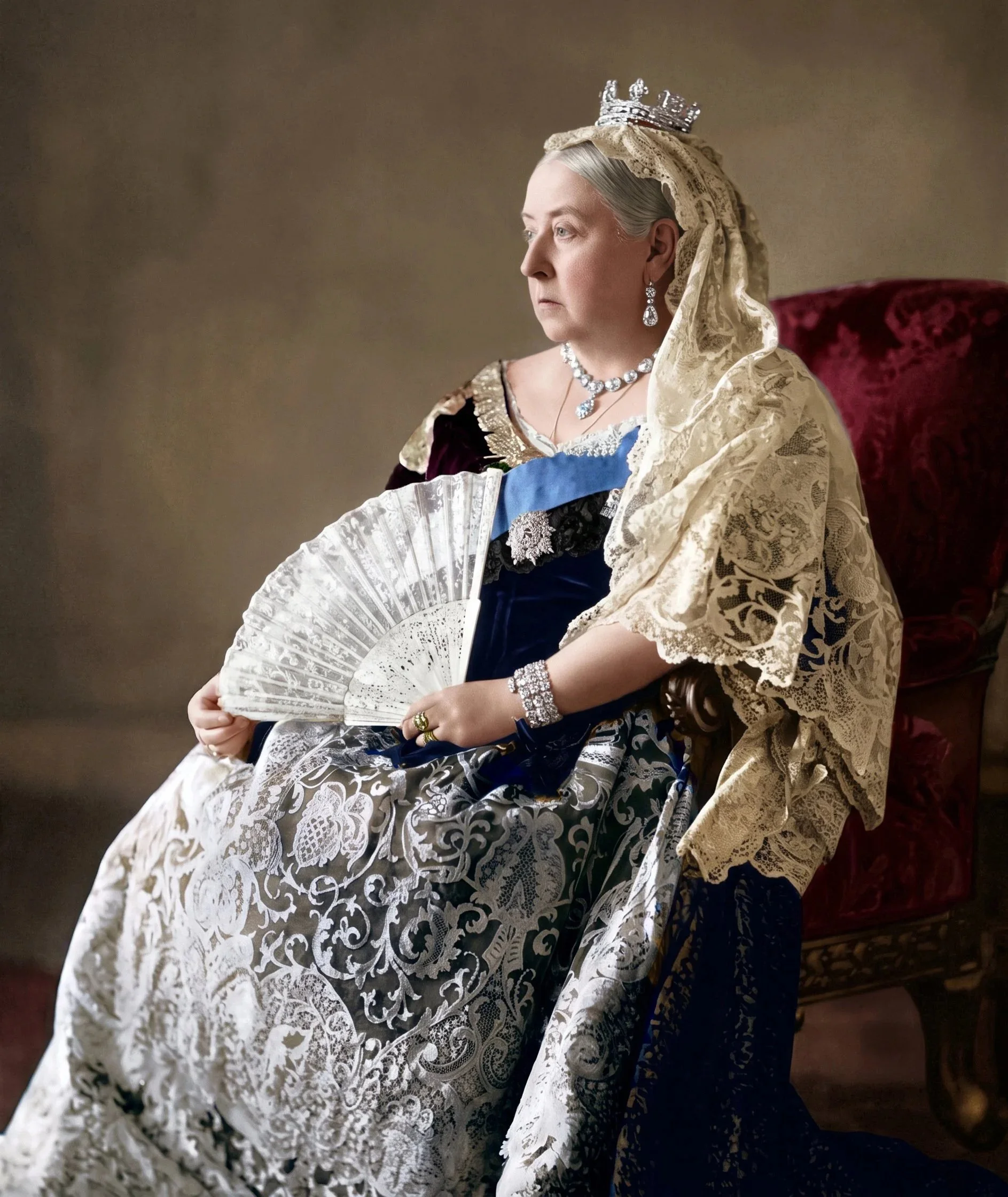 Colourised photo of Queen Victoria dressed in elaborate, historical attire with a crown, lace shawl, and jewellery, sitting on a red velvet chair, holding a decorative fan.