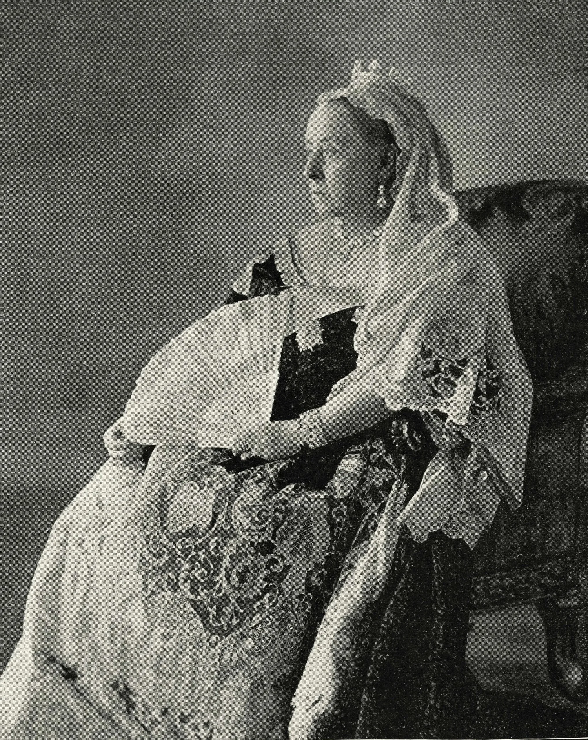 Black and white photo of Queen Victoria dressed in elaborate, historical attire with a crown, lace shawl, and jewellery,