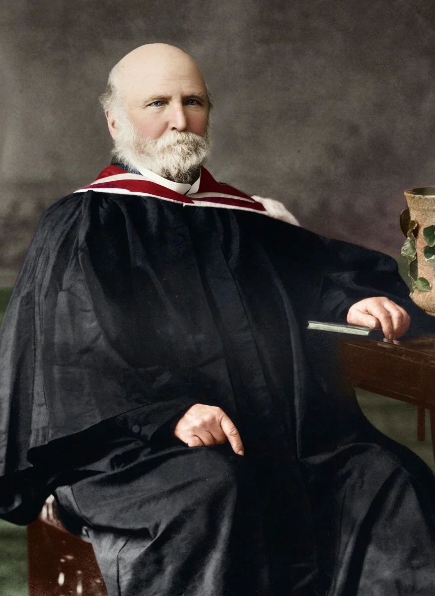 A distinguished older man with a beard and mustache dressed in academic regalia, sitting with one hand resting on a wooden desk.