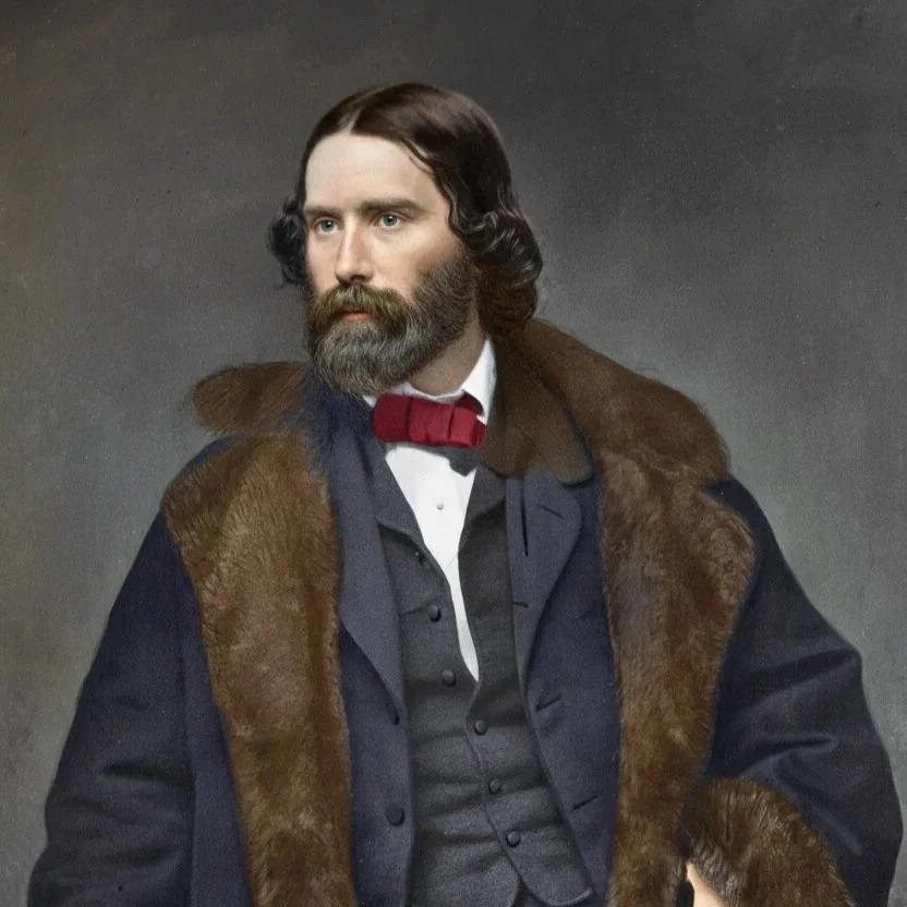 A man with long, dark, wavy hair and a full beard wearing a dark coat with a fur collar, a dark vest, a white shirt, and a red bow tie, posed against a plain background.