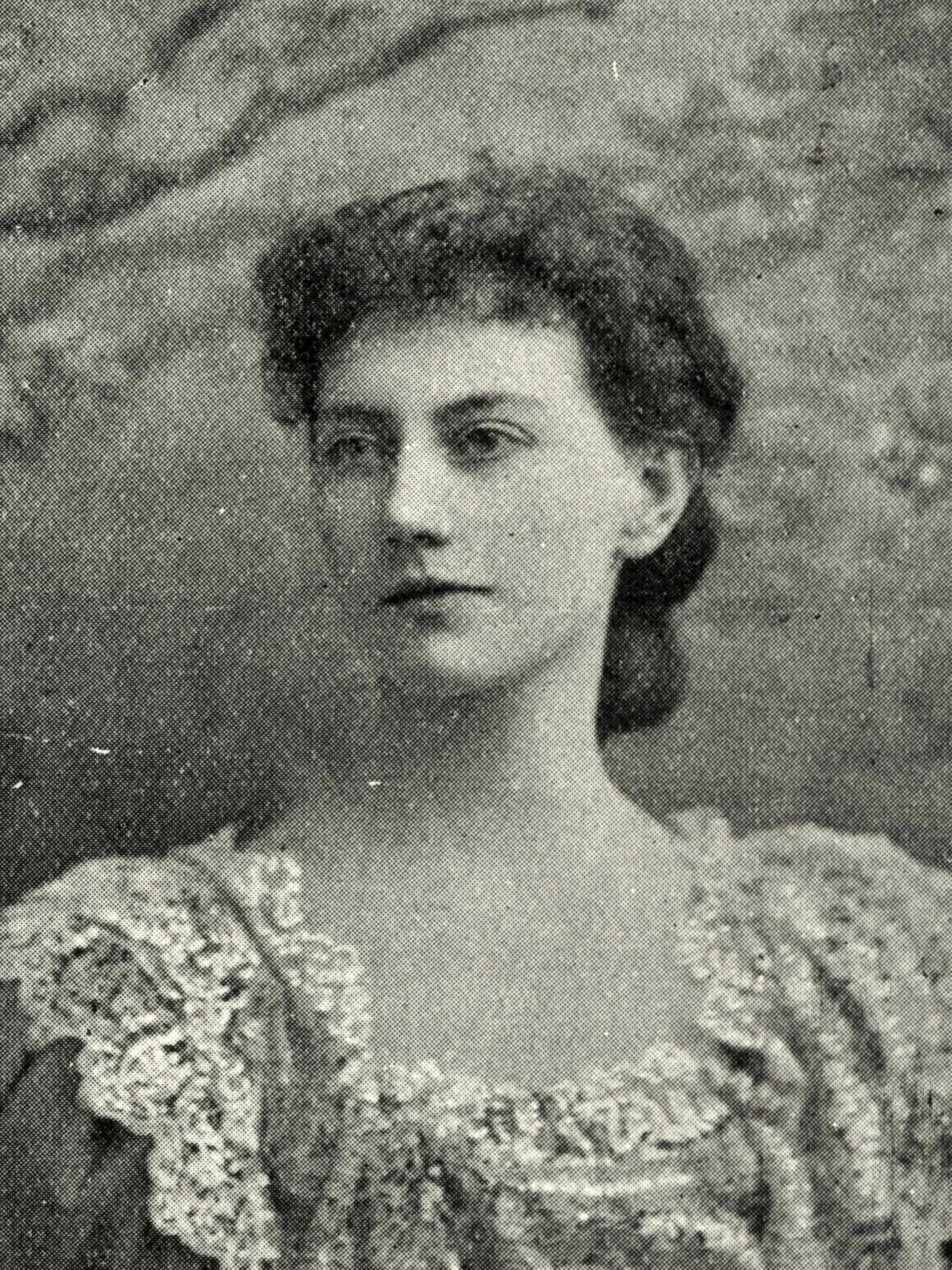 A black and white photo of a woman with dark, short, wavy hair, wearing a patterned dress with ornate detailing on the collar and sleeves.