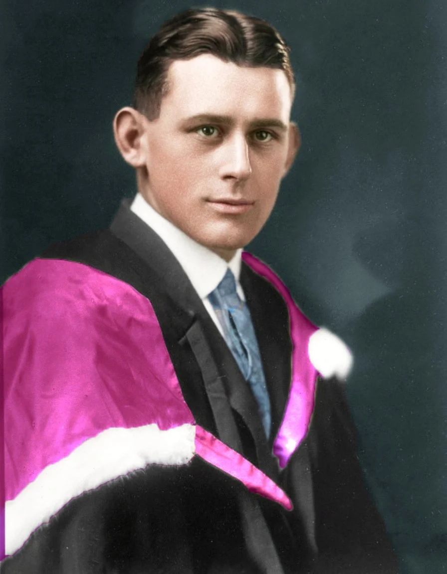 A young man in a suit, wearing a black gown with magenta and white accents, likely a graduation robe, against a dark background.