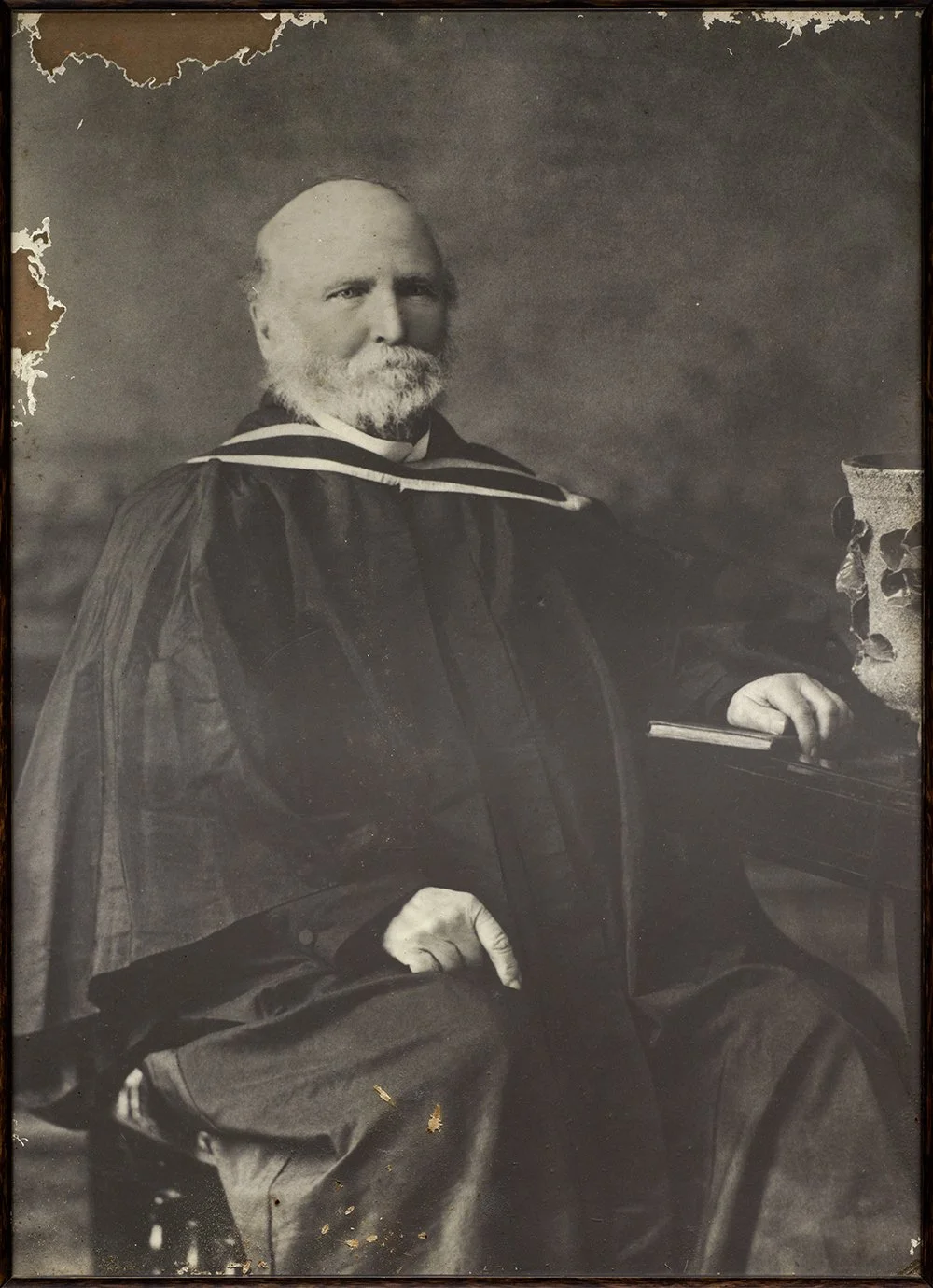 Old black-and-white portrait of a man wearing academic robes with a beard and mustache, seated next to a bust of a woman