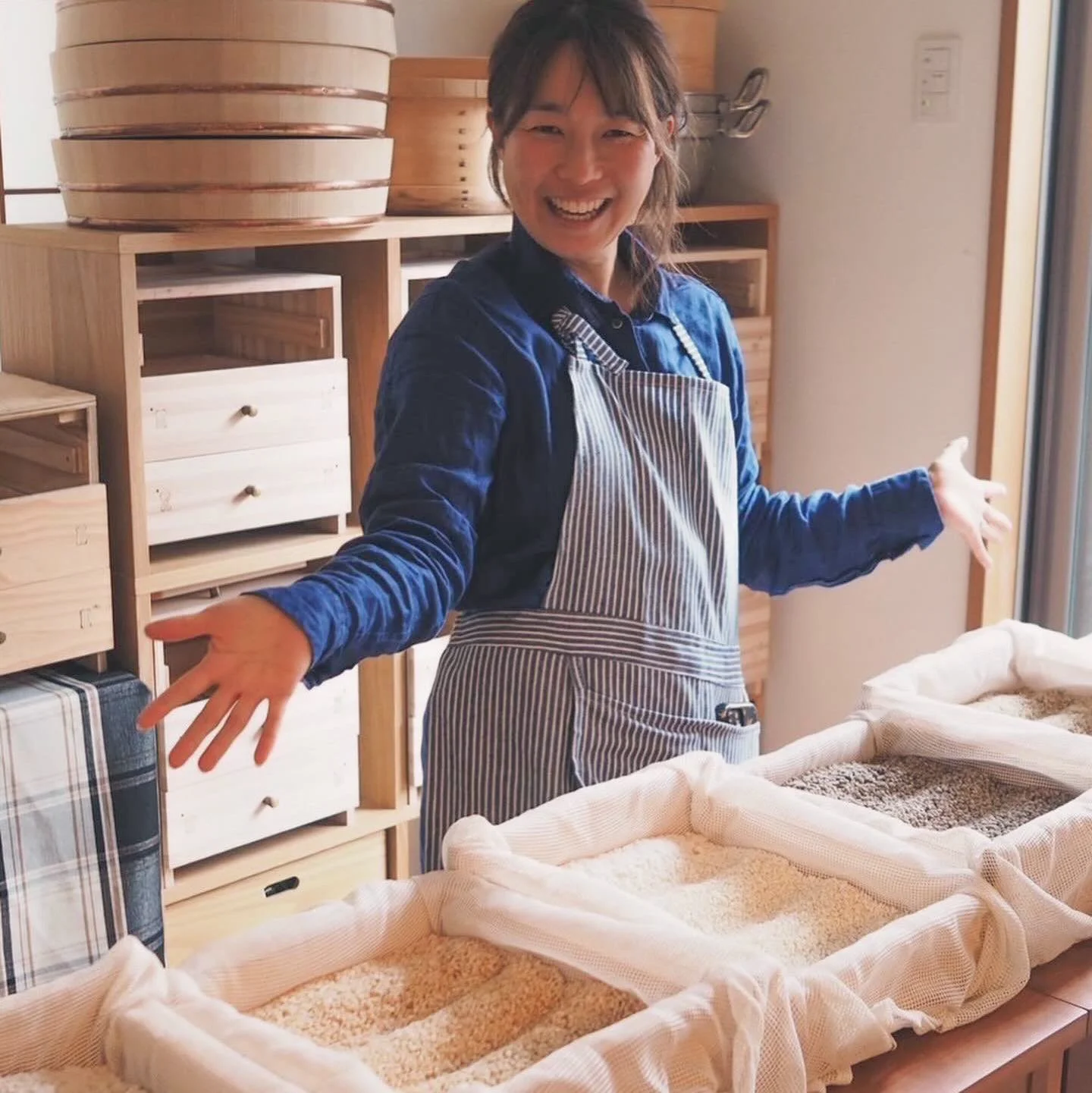 A Japanese woman standing in a kitchen with baskets of rice on the bench in front of her