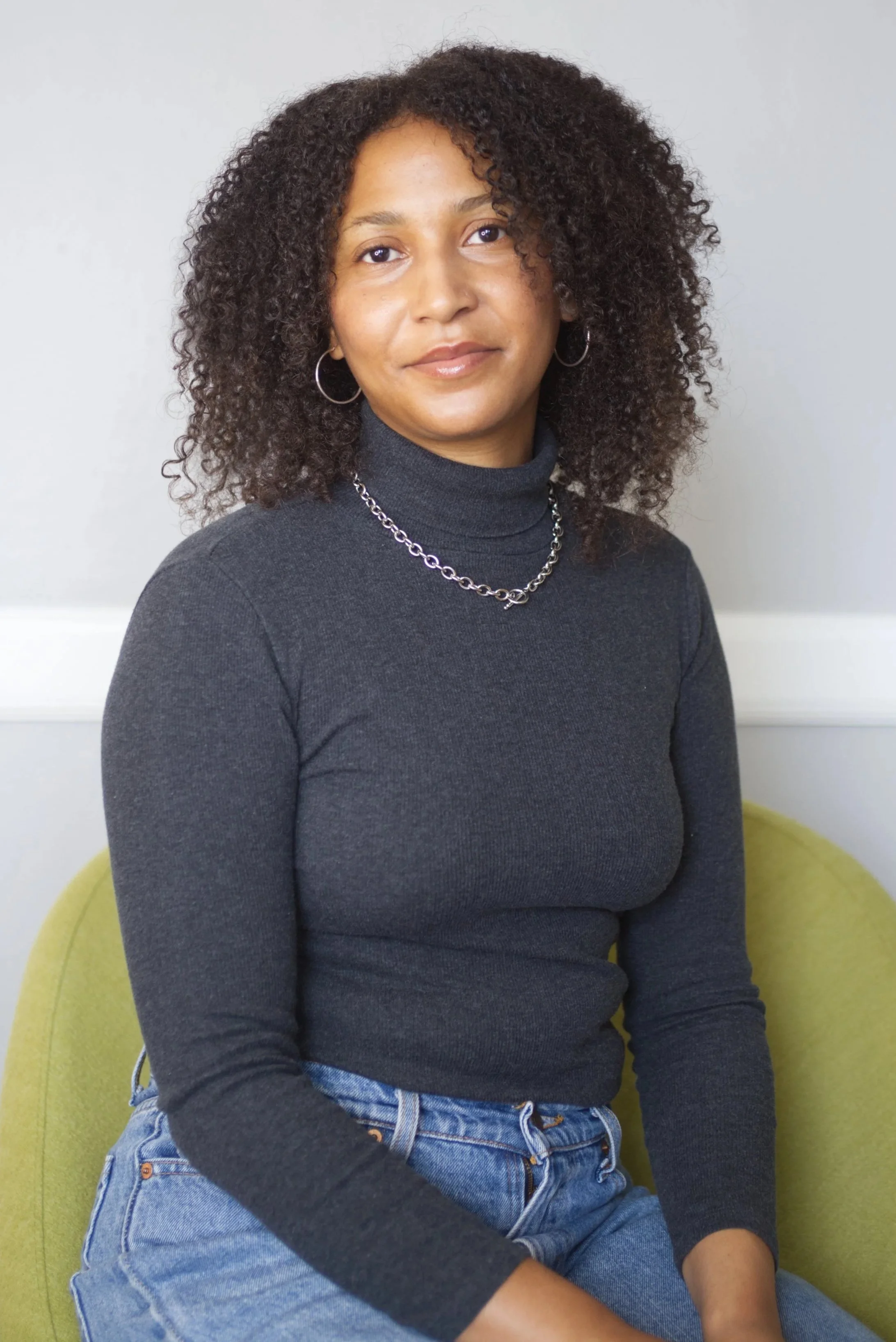 A woman sitting in a yellow chair with a light grey background, wearing a black turtleneck, silver chain necklace, hoop earrings, and blue jeans.