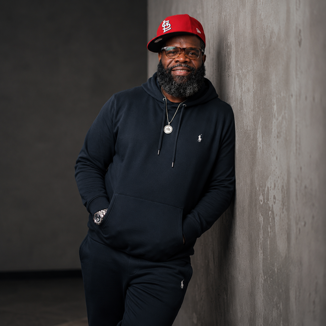 Man with glasses and beard wearing a black hoodie, red cap, and jewelry, leaning against a concrete wall.