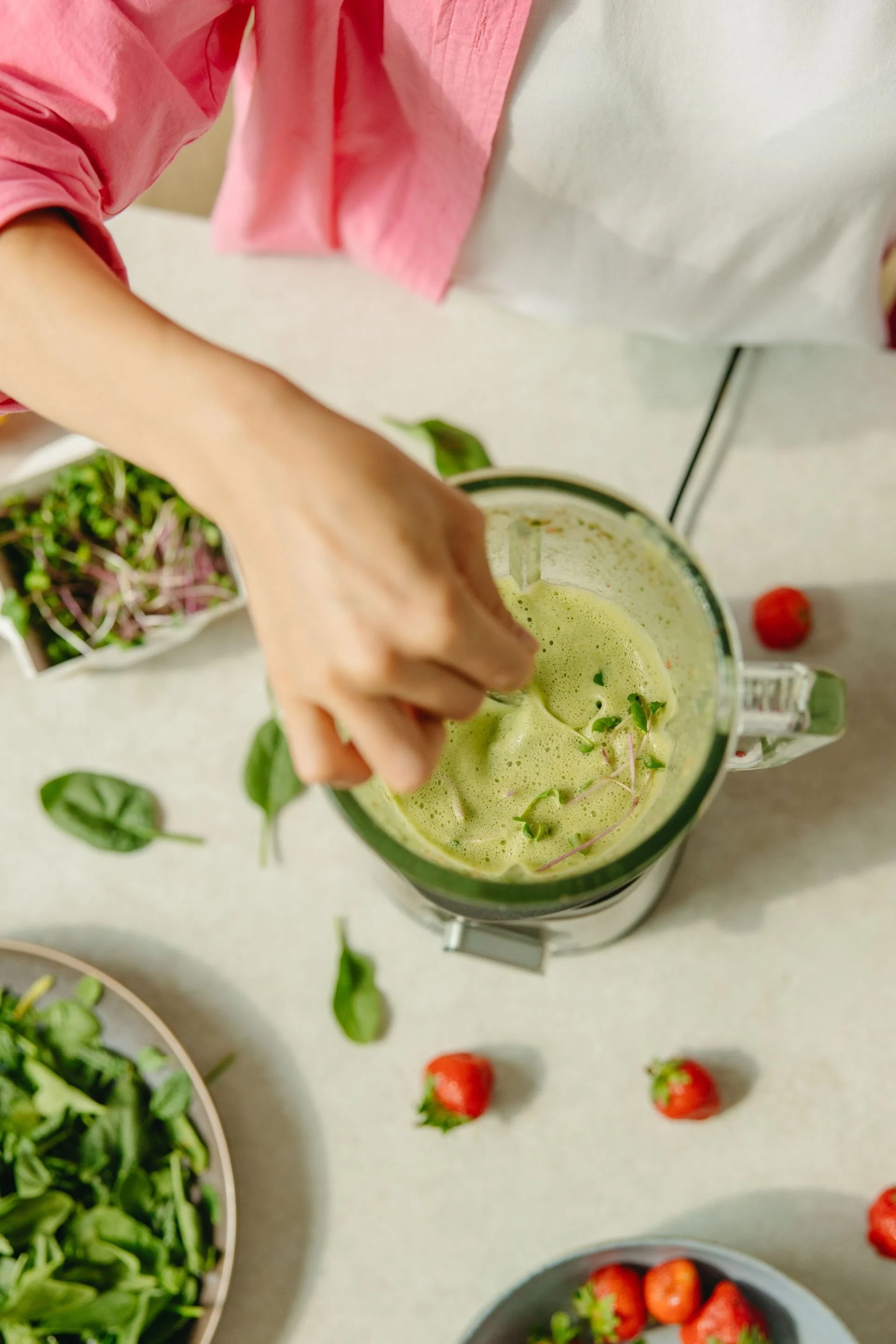 Close-up of a person blending green smoothie with leafy greens and microgreens, assorted strawberries, cherry tomatoes, and other vegetables on a white surface.