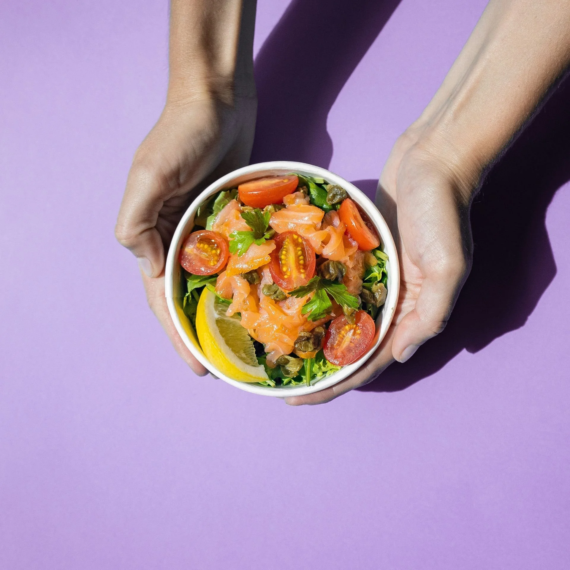 A bowl of salad with cherry tomatoes, smoked salmon, capers, and garnished with parsley, held by a person with light skin against a purple background.