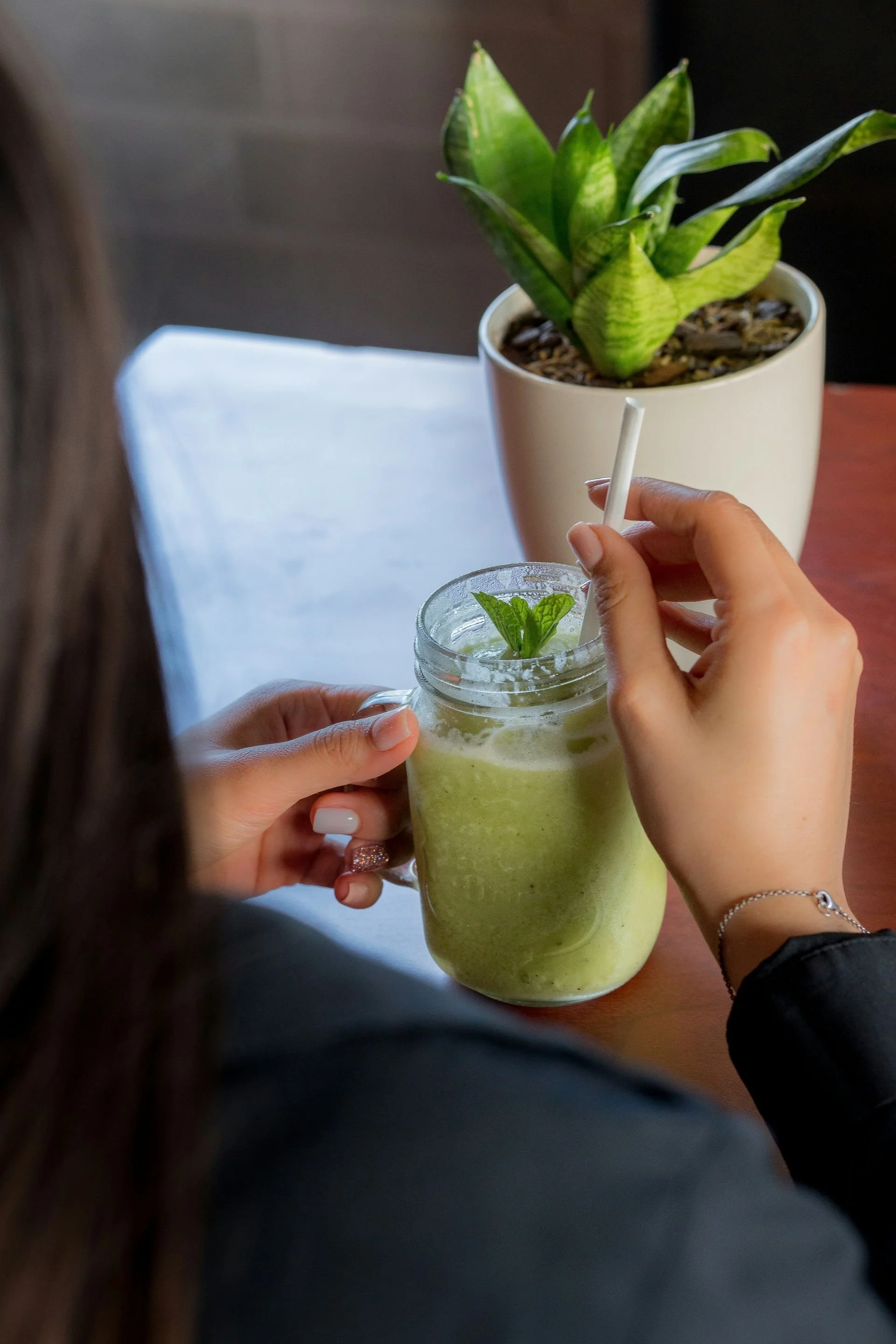 Person holding a glass of green smoothie with mint leaves, with a potted green plant in the background.