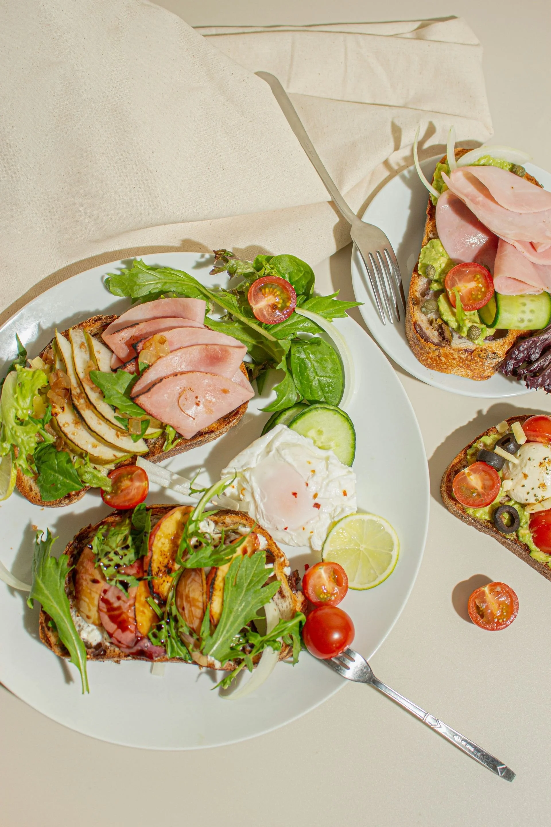 Close-up of open-faced sandwiches with various toppings, cherry tomatoes, cucumber slices, lemon wedge, and a poached egg on a white plate, with a fork nearby.